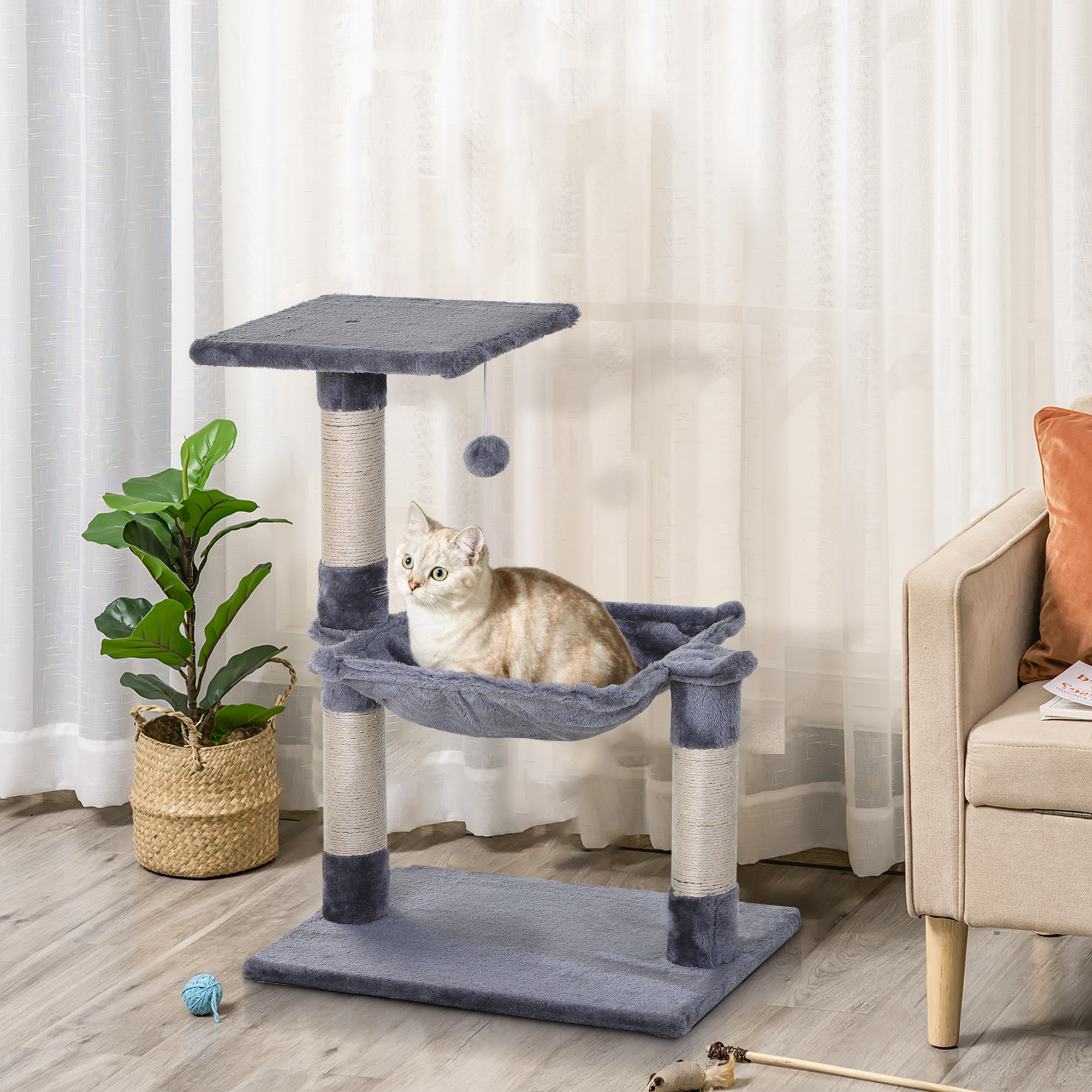 Un gato en un árbol para gatos gris con una hamaca y un juguete. Ventana y cortinas en el fondo.