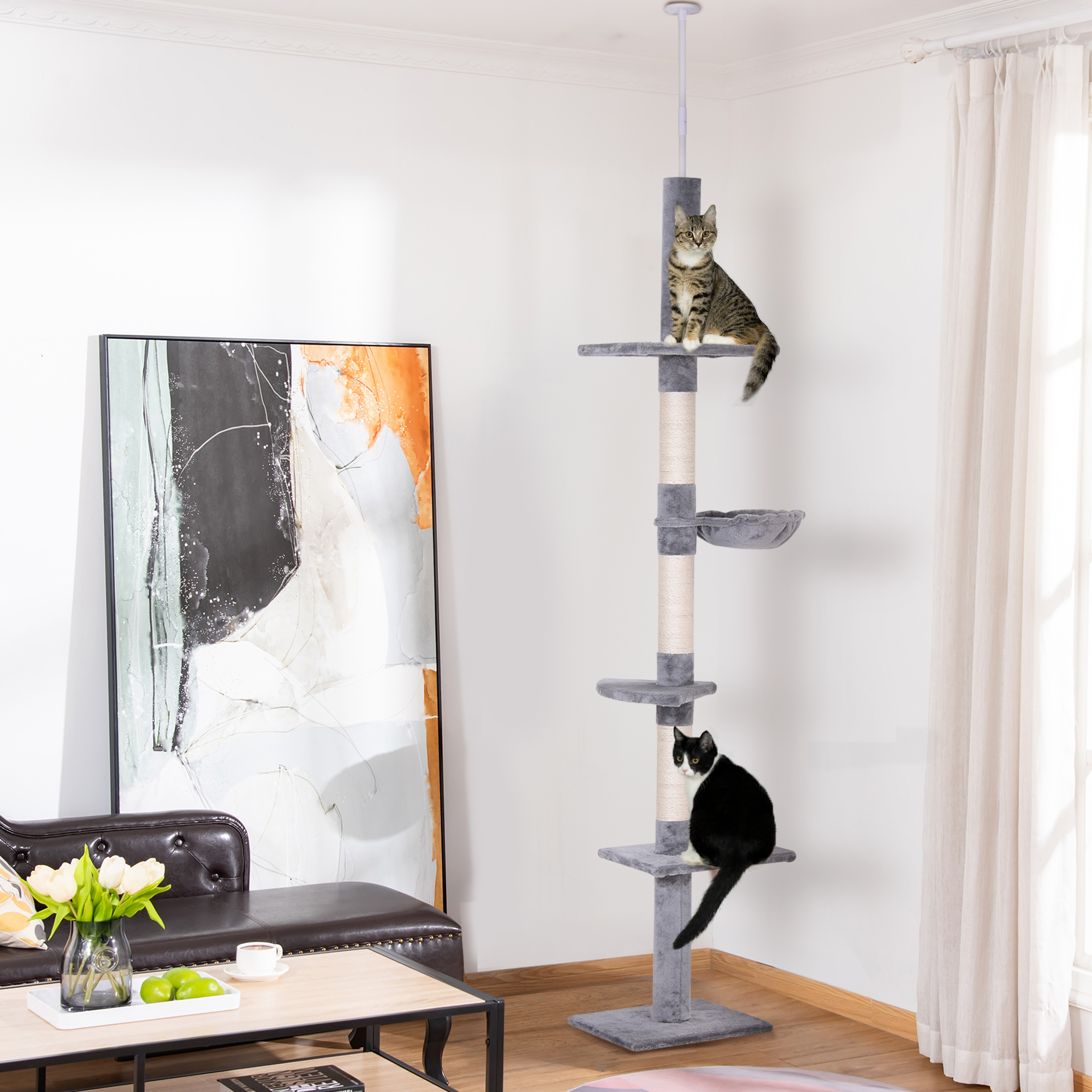 Dos gatos en un árbol para gatos alto. El árbol tiene plataformas, una cama y postes para rascar. Ventana y arte en el fondo.