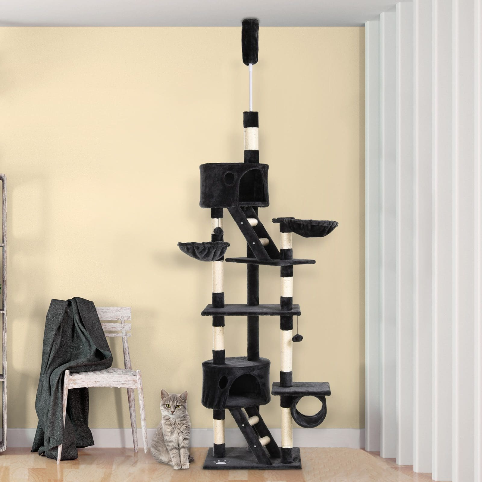 Un árbol para gatos grande negro y beige con múltiples niveles, un gato sentado en la base y una silla en el fondo.