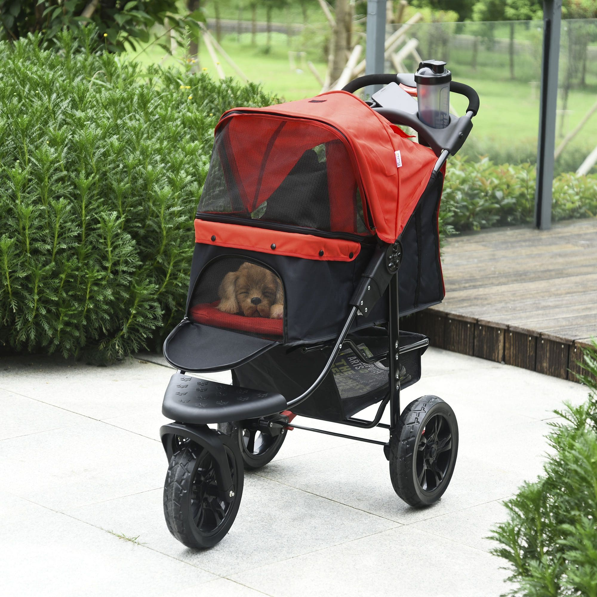 Un cochecito rojo y negro para mascotas con un perro adentro. El cochecito está sobre una superficie pavimentada rodeada de vegetación.