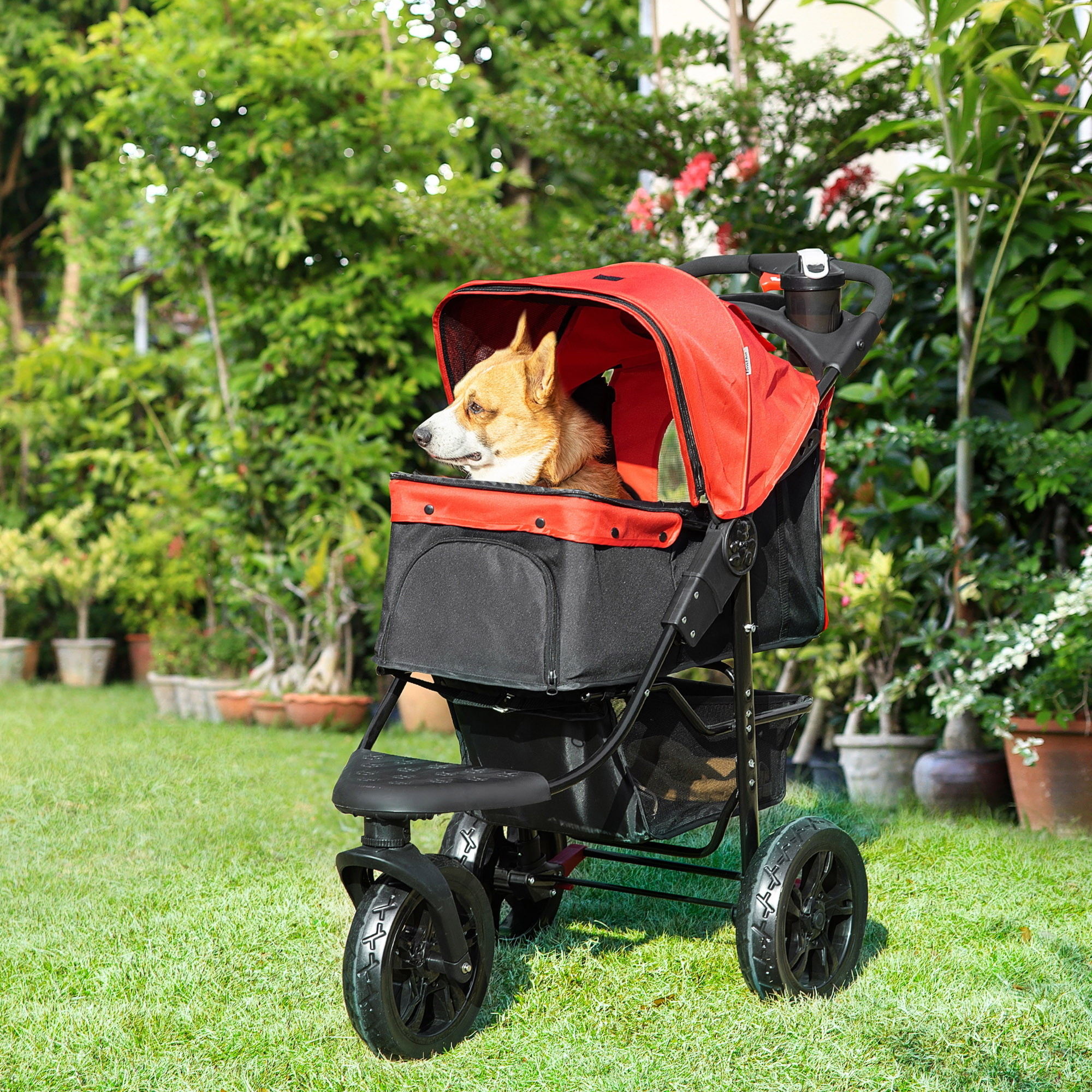 Un perro en un cochecito rojo para mascotas en un césped verde, rodeado de plantas. El cochecito tiene detalles negros.