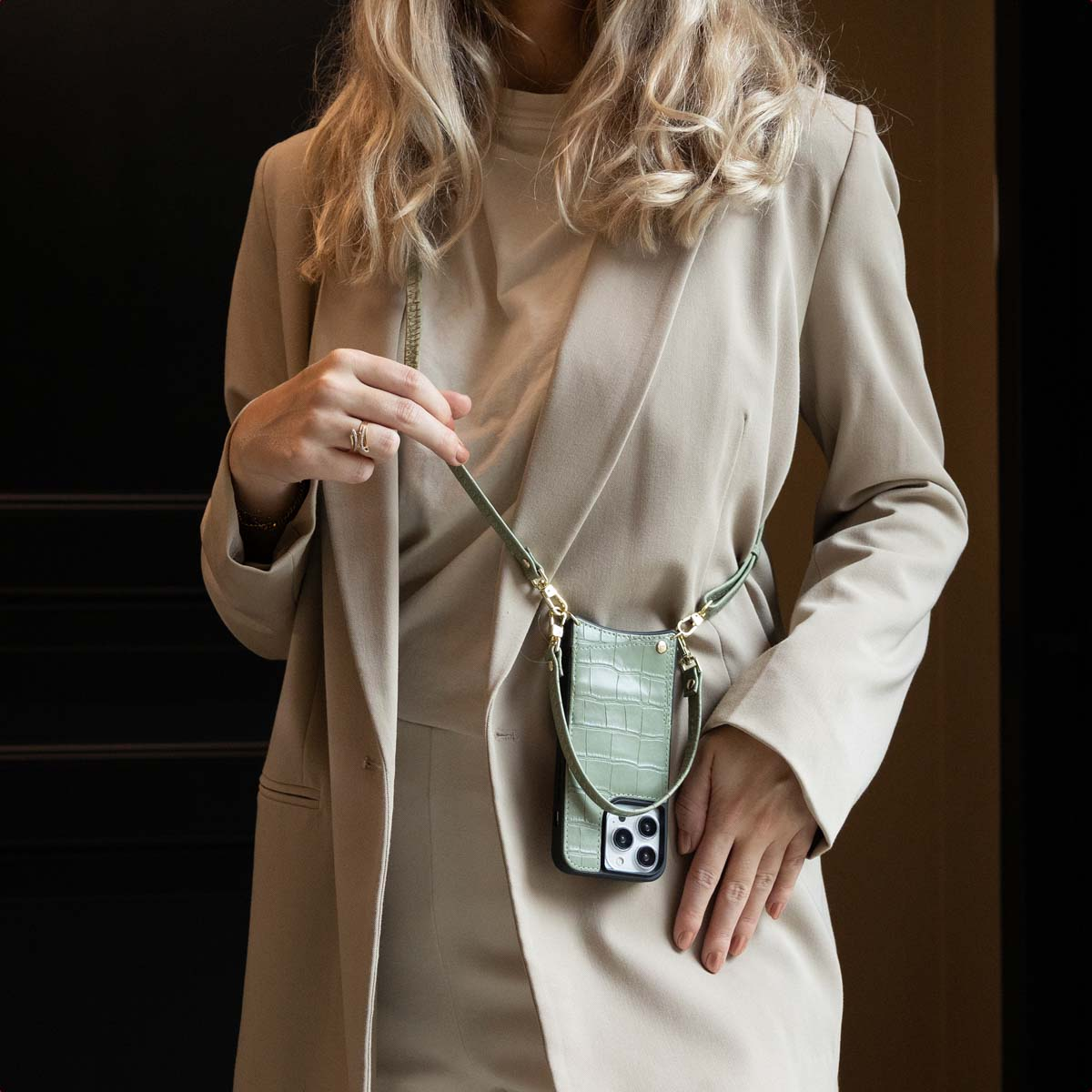 Een vrouw in een beige blazer en jurk houdt een groene telefoonhoes met een band vast. De telefoonhoes heeft een krokodillenpatroon.