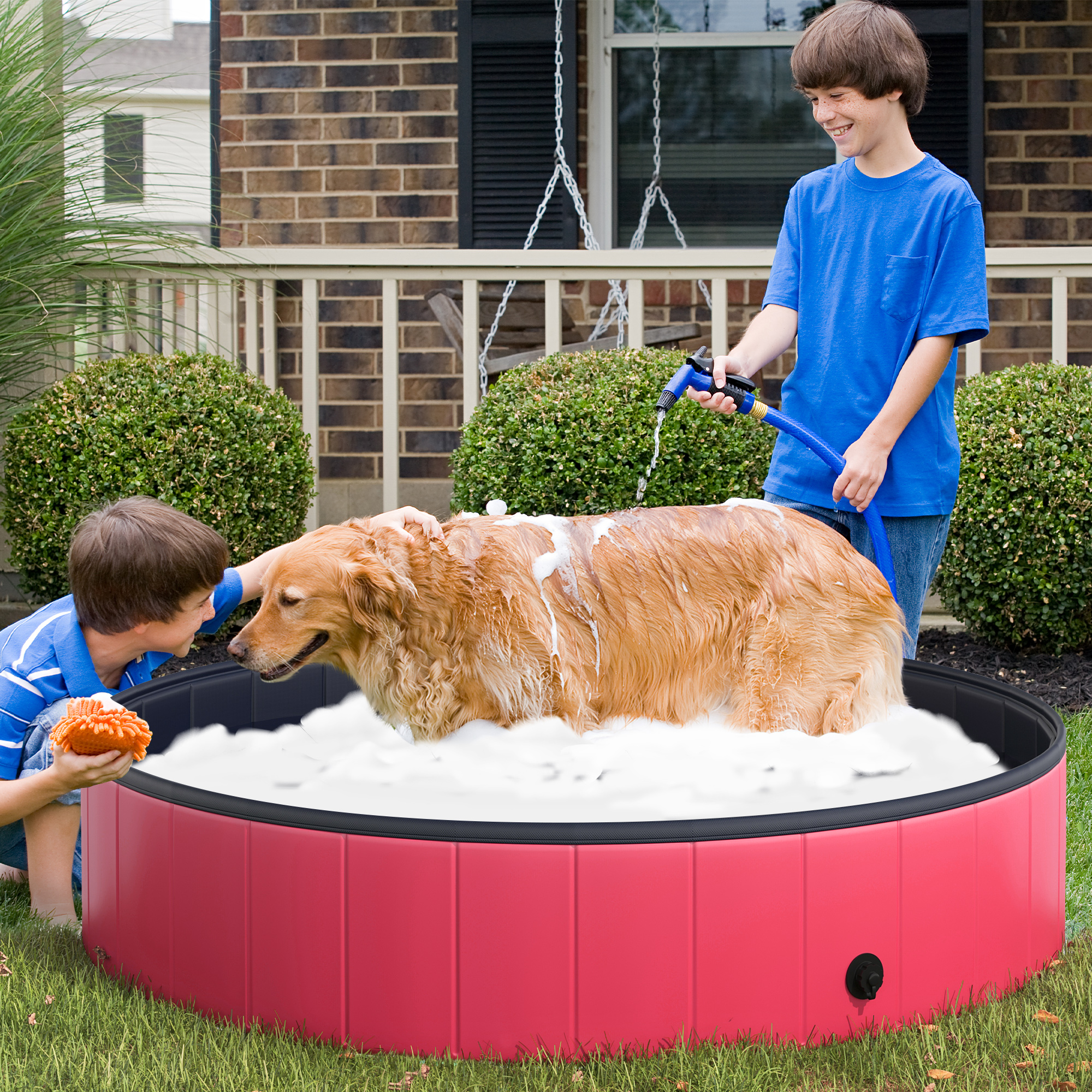 Dos niños lavan a un perro en una piscina de plástico roja. El perro está cubierto de espuma de jabón.