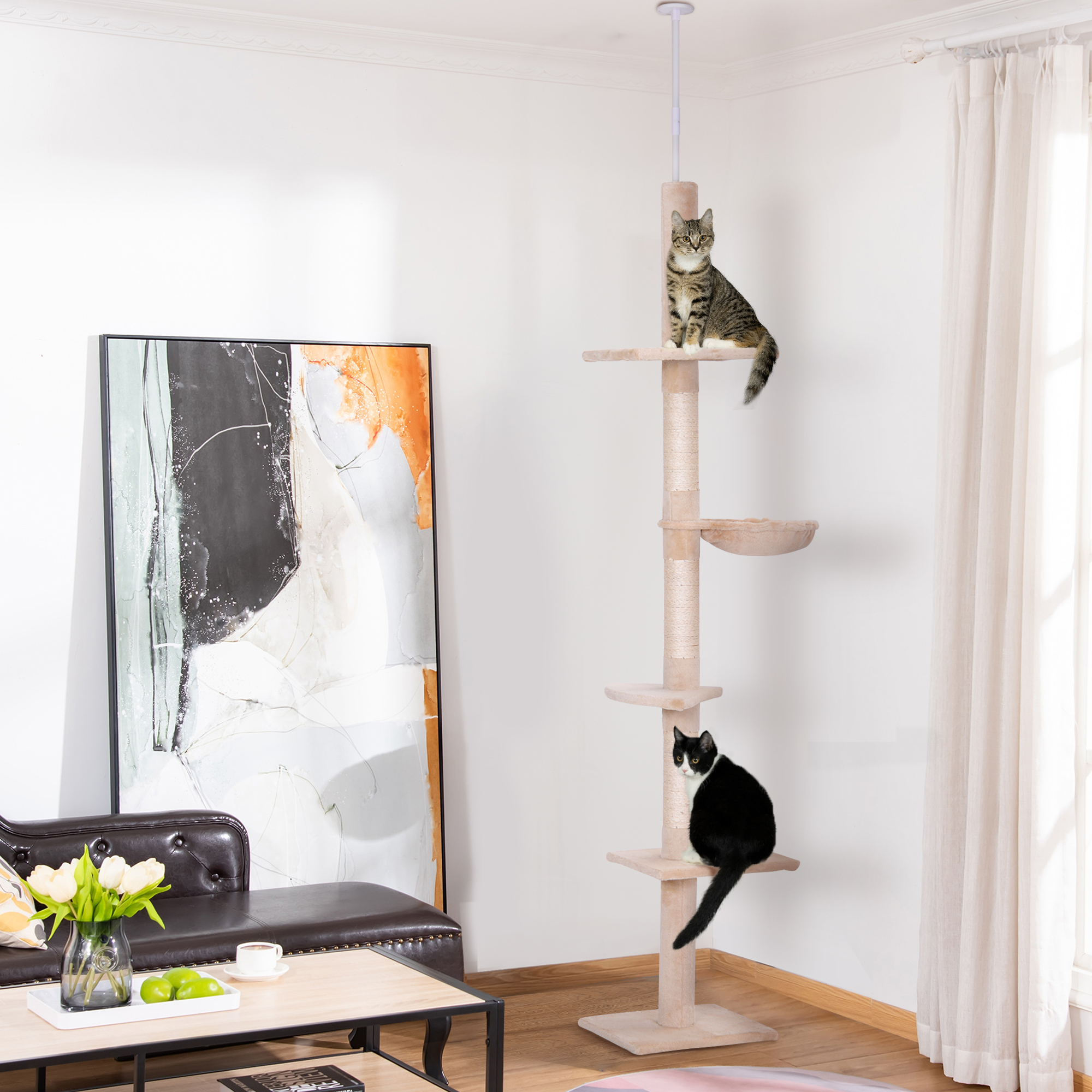 Dos gatos en un árbol para gatos beige. Uno blanco y negro, el otro atigrado. Habitación con arte y sofá.