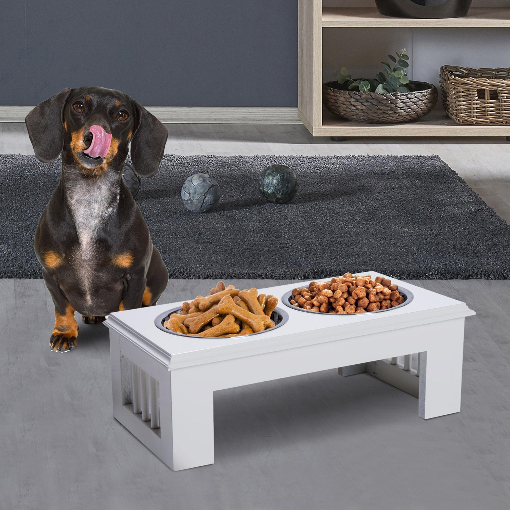 Un perro lamiéndose la nariz junto a un soporte blanco para comida con dos cuencos.