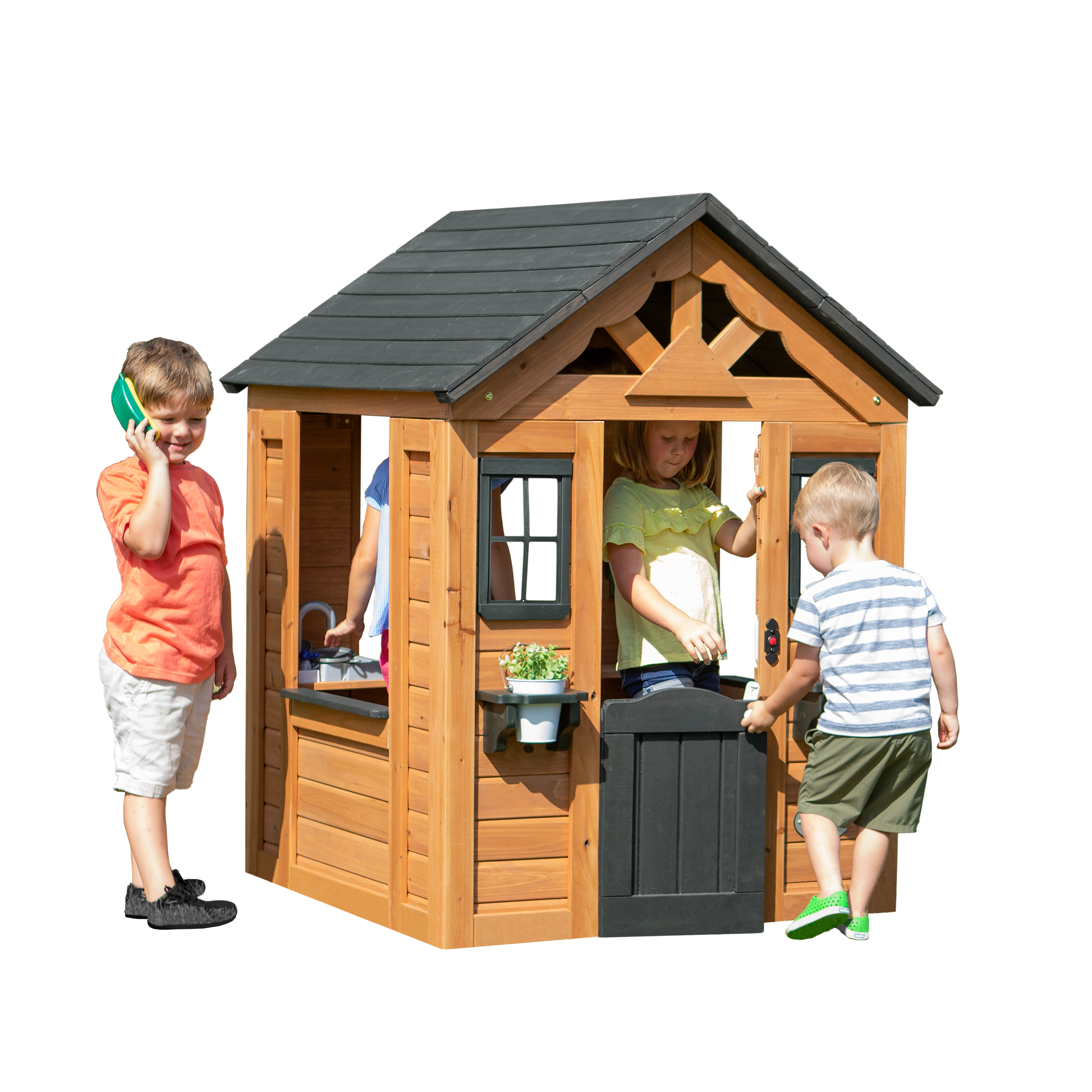 Kinder spielen in einem Holzspielhaus. Zwei Kinder sind drinnen, eines draußen mit einem Telefon.