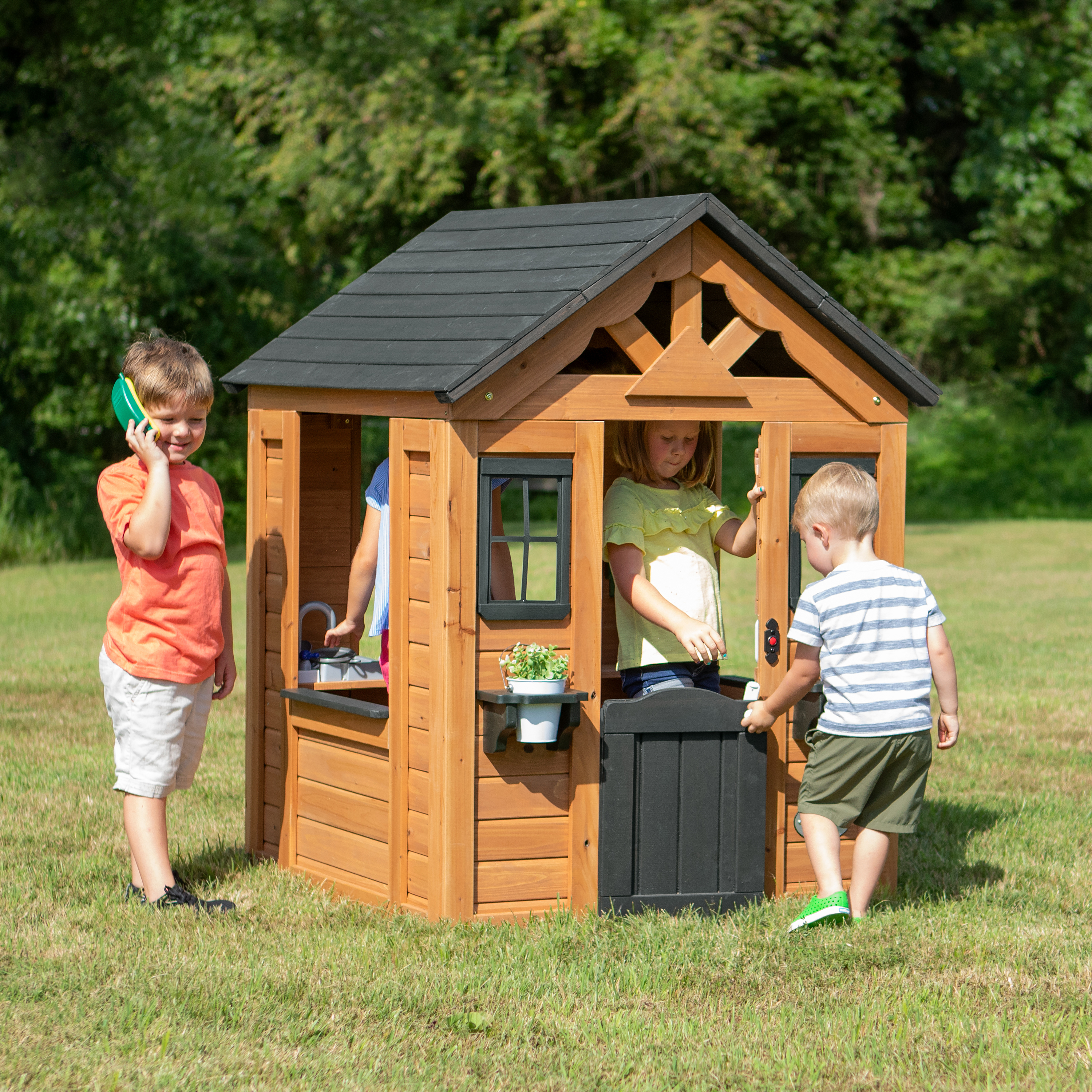 Kinder spielen in einem hölzernen Spielhaus. Das Haus hat eine Tür, Fenster und ein schwarzes Dach.