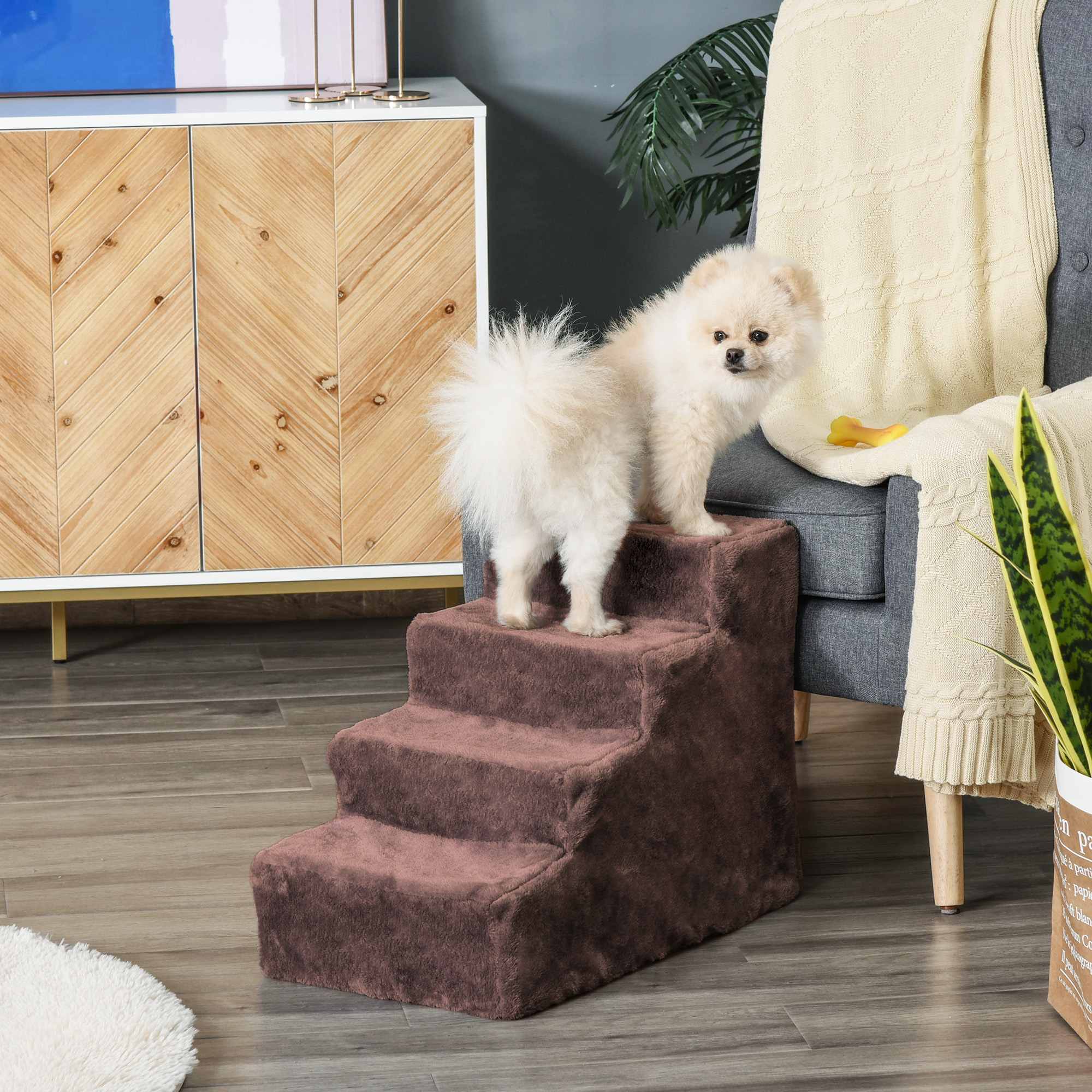 Perro pequeño de pie en escalones marrones para mascotas, con una silla gris y otros objetos en el fondo.