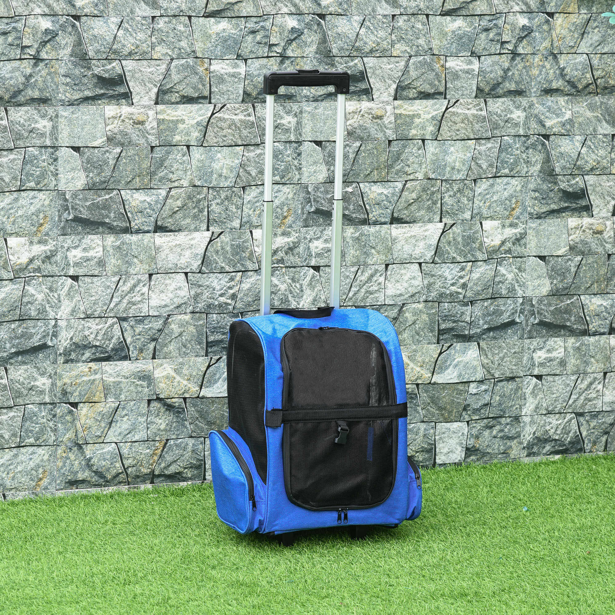 Un transportín azul para mascotas sobre ruedas con un asa plateada se encuentra sobre césped verde contra una pared de piedra.