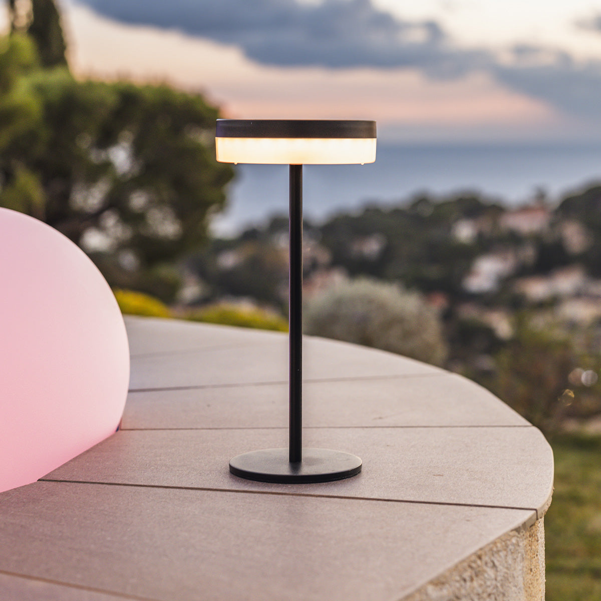Schwarze Außenlampe auf grauer Oberfläche, mit einem rosa Objekt im Hintergrund.