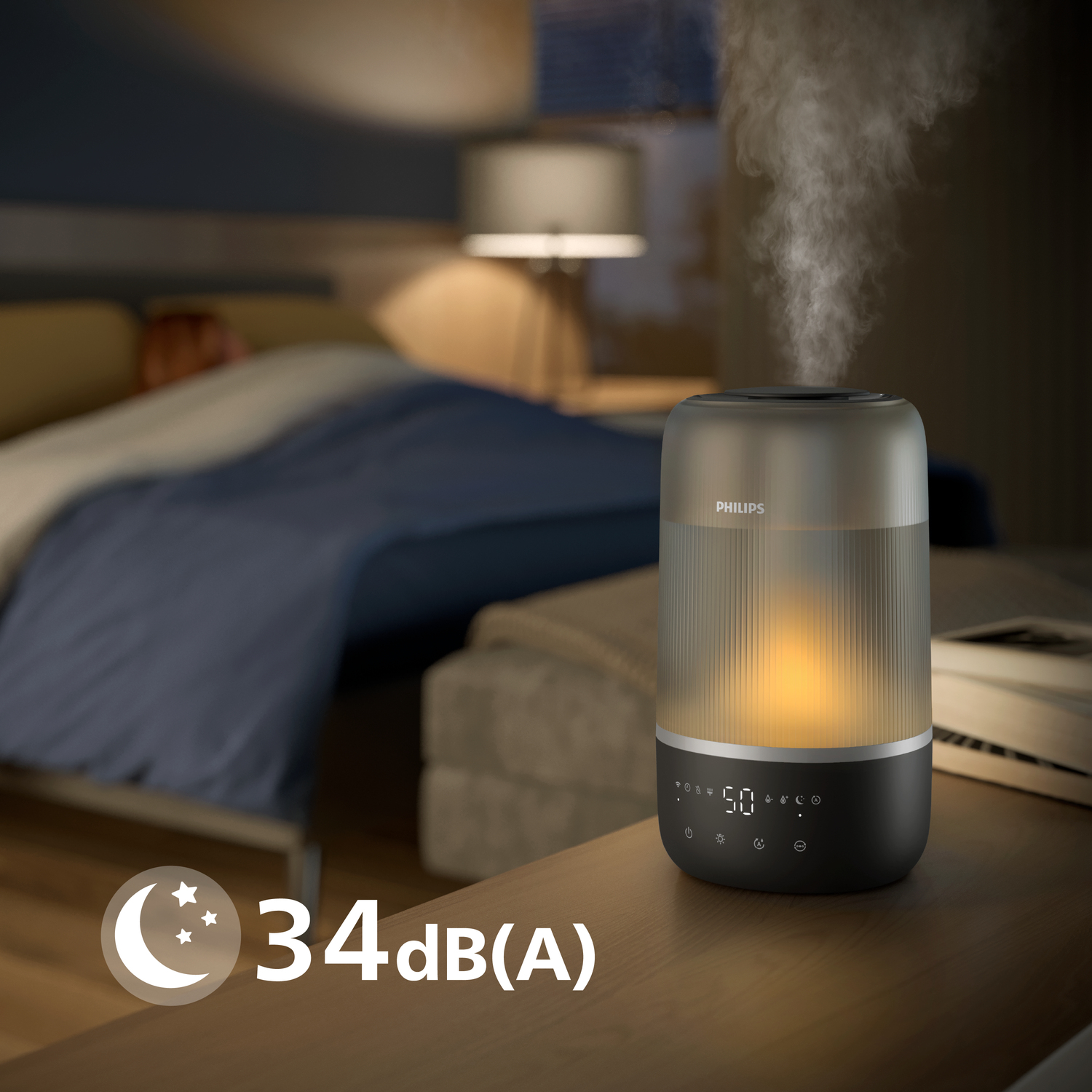 Nawilżacz stoi na stole. Para z niego unosi się. Łóżko jest w rozmytym tle.