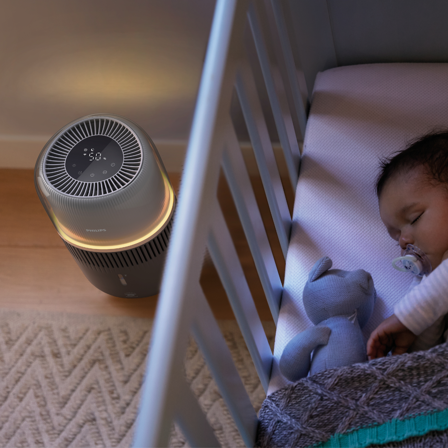 Ein Baby schläft in einem Kinderbett mit einem Luftreiniger in der Nähe. Warmes Licht und Teddybär.