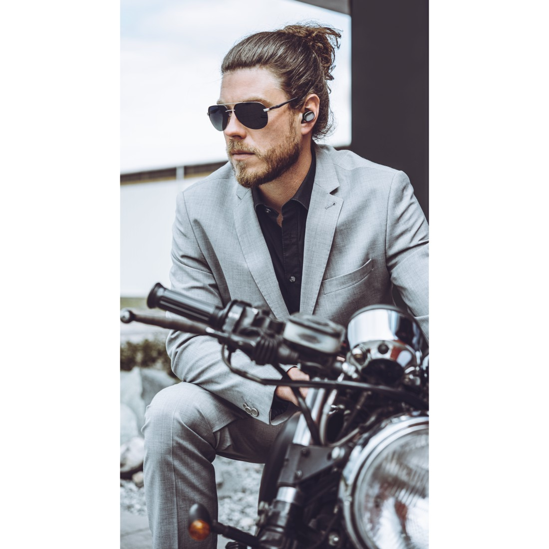 Mężczyzna w szarym garniturze i okularach przeciwsłonecznych siedzi na motocyklu, nosząc słuchawki.