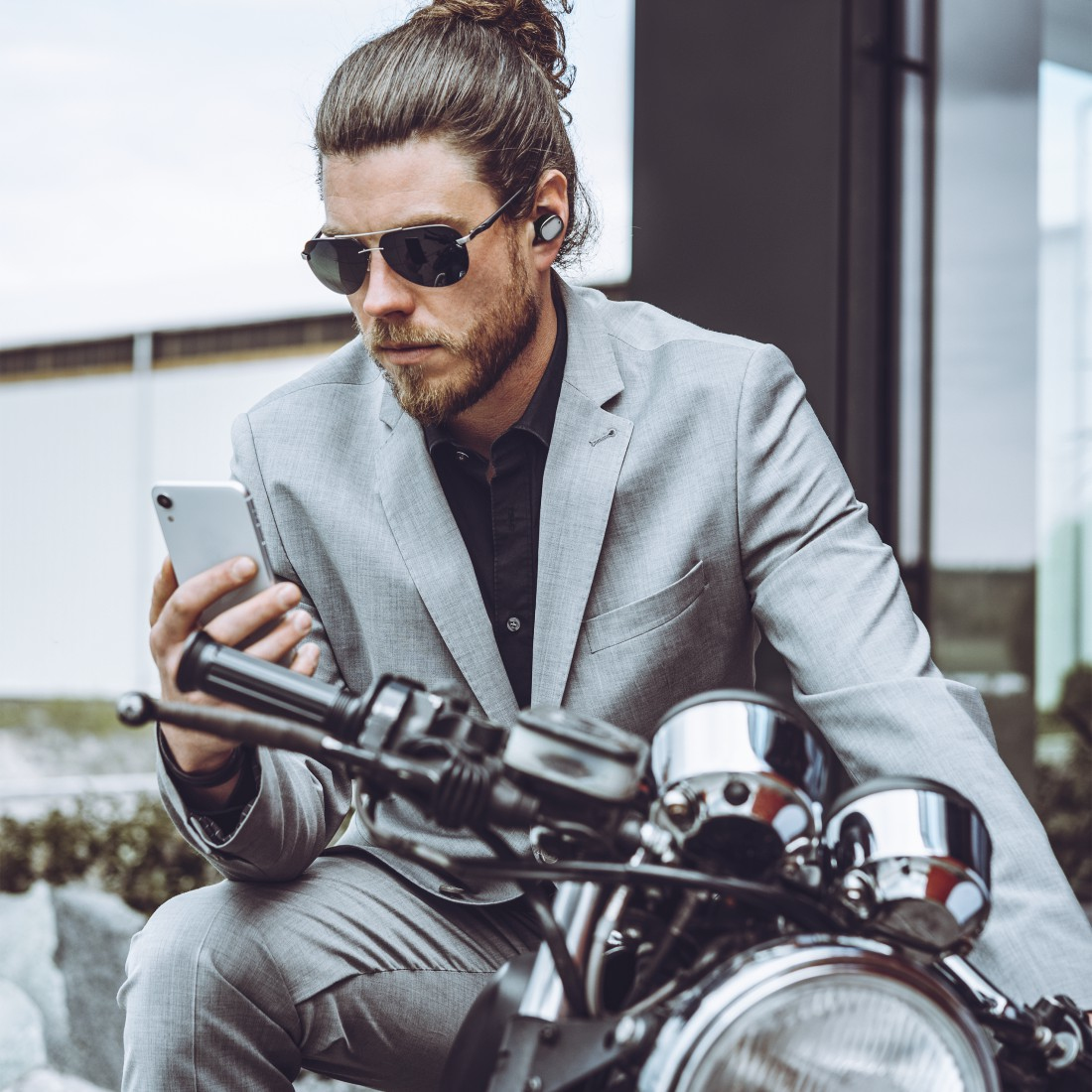 Mężczyzna w okularach przeciwsłonecznych i słuchawkach, patrzący na swój telefon siedząc na motocyklu.