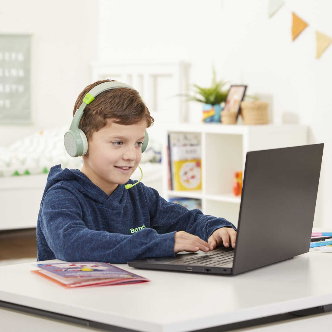 Ein Junge mit Kopfhörern benutzt einen Laptop. Auf dem Schreibtisch liegt ein Notizbuch.