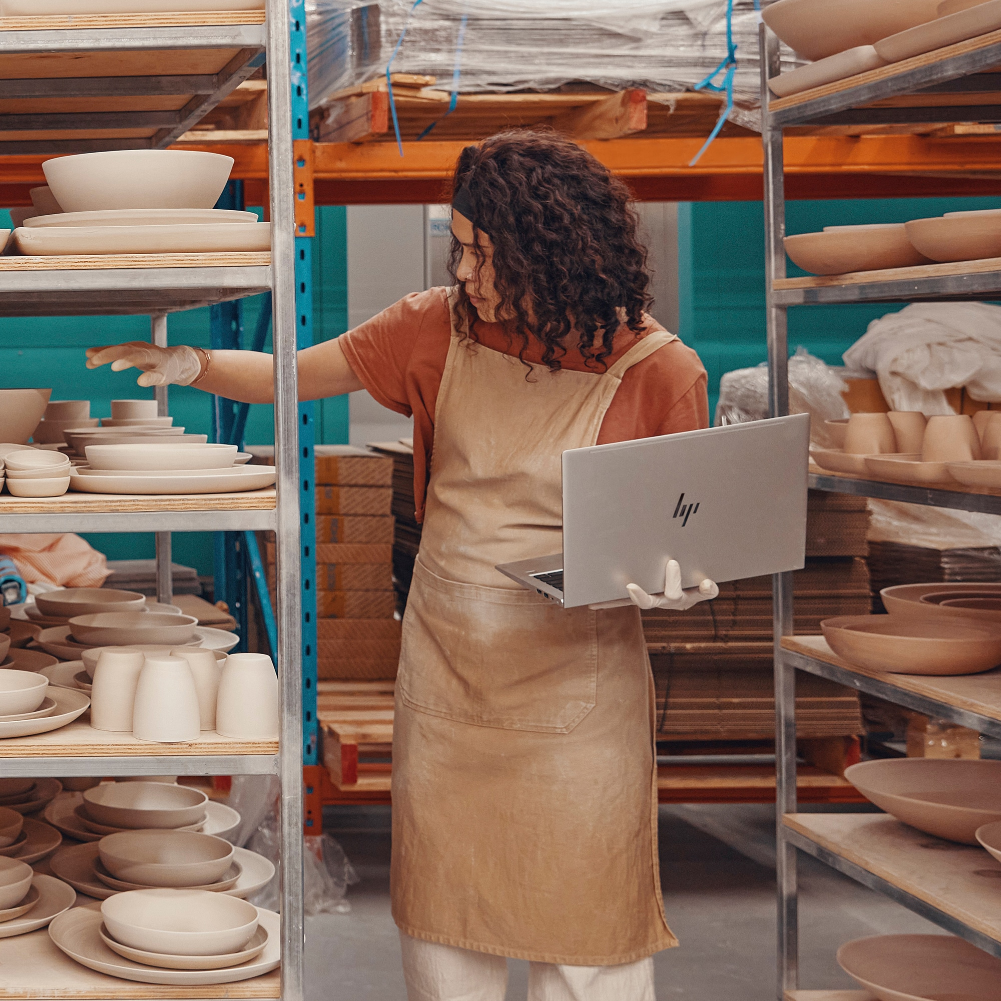 Una donna in uno studio di ceramica con un laptop, che guarda scaffali di oggetti in ceramica.