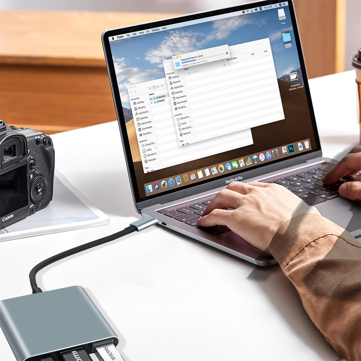 Eine Person benutzt einen Laptop, der mit einem USB-Hub verbunden ist, und eine Canon-Kamera ist sichtbar.