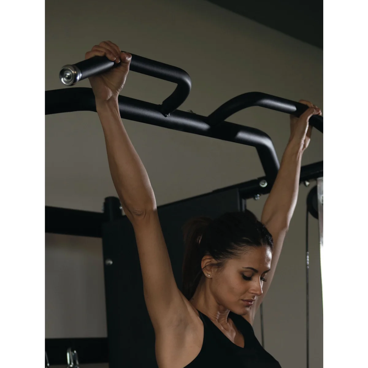 Een vrouw die pull-ups doet op een zwarte gymbar. Ze draagt een zwarte top. De achtergrond is neutraal.
