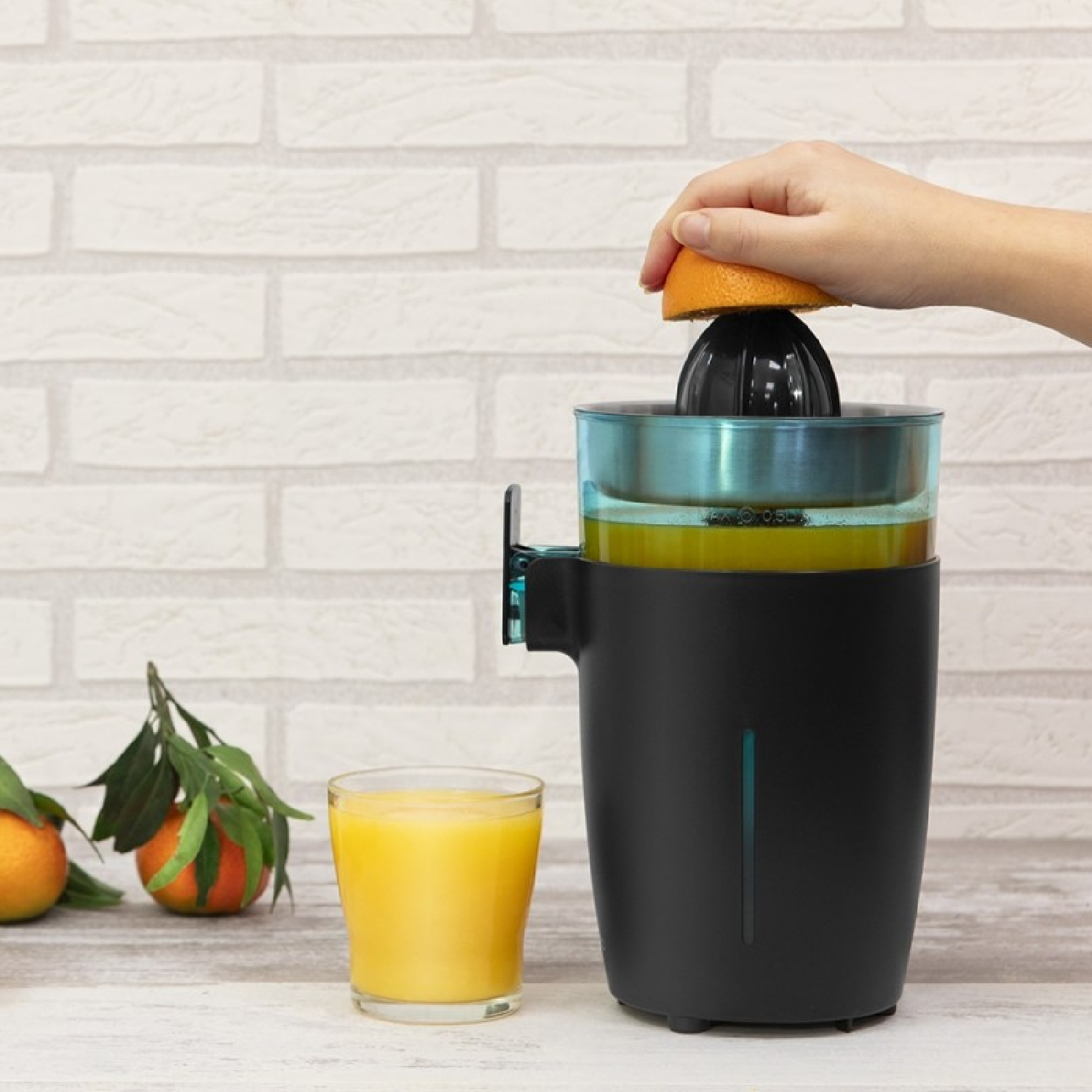 Persona exprimiendo media naranja en una exprimidora. Vaso de zumo y naranjas en la encimera.