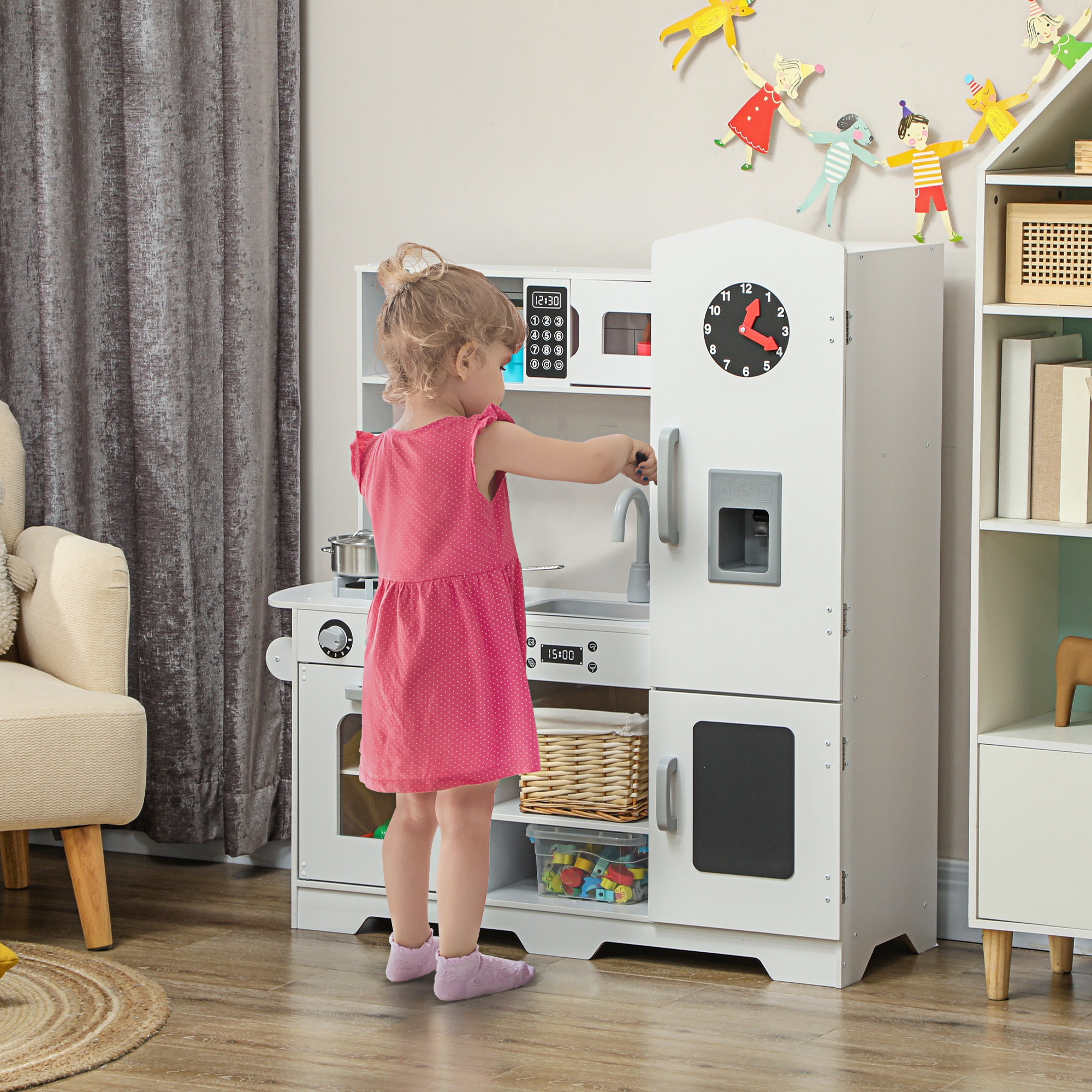 Una niña con un vestido rosa en una cocina de juguete blanca. Se ve un reloj, microondas y nevera.