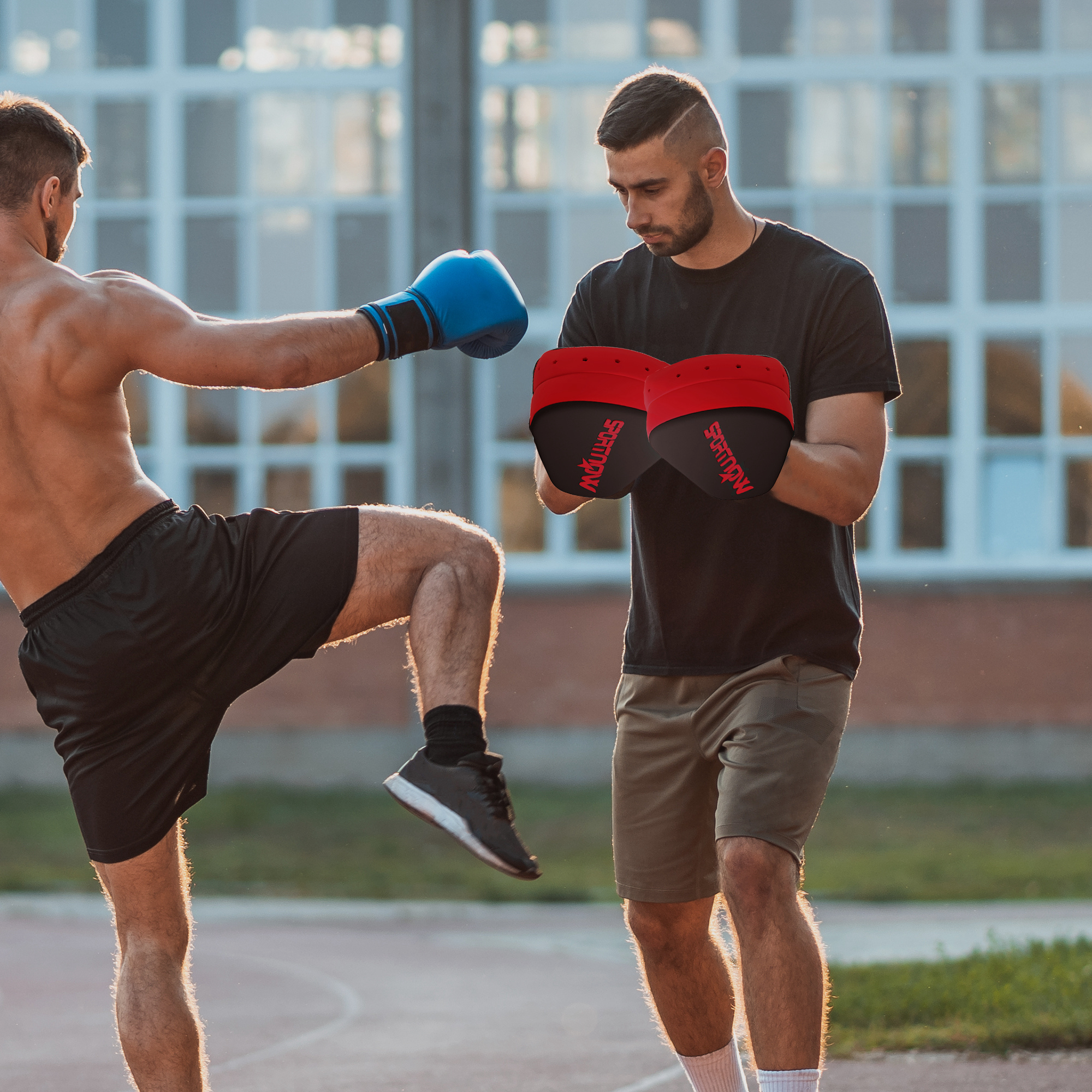 Dos hombres boxeando. Uno patea, guantes azules, el otro sostiene manoplas, guantes rojos.