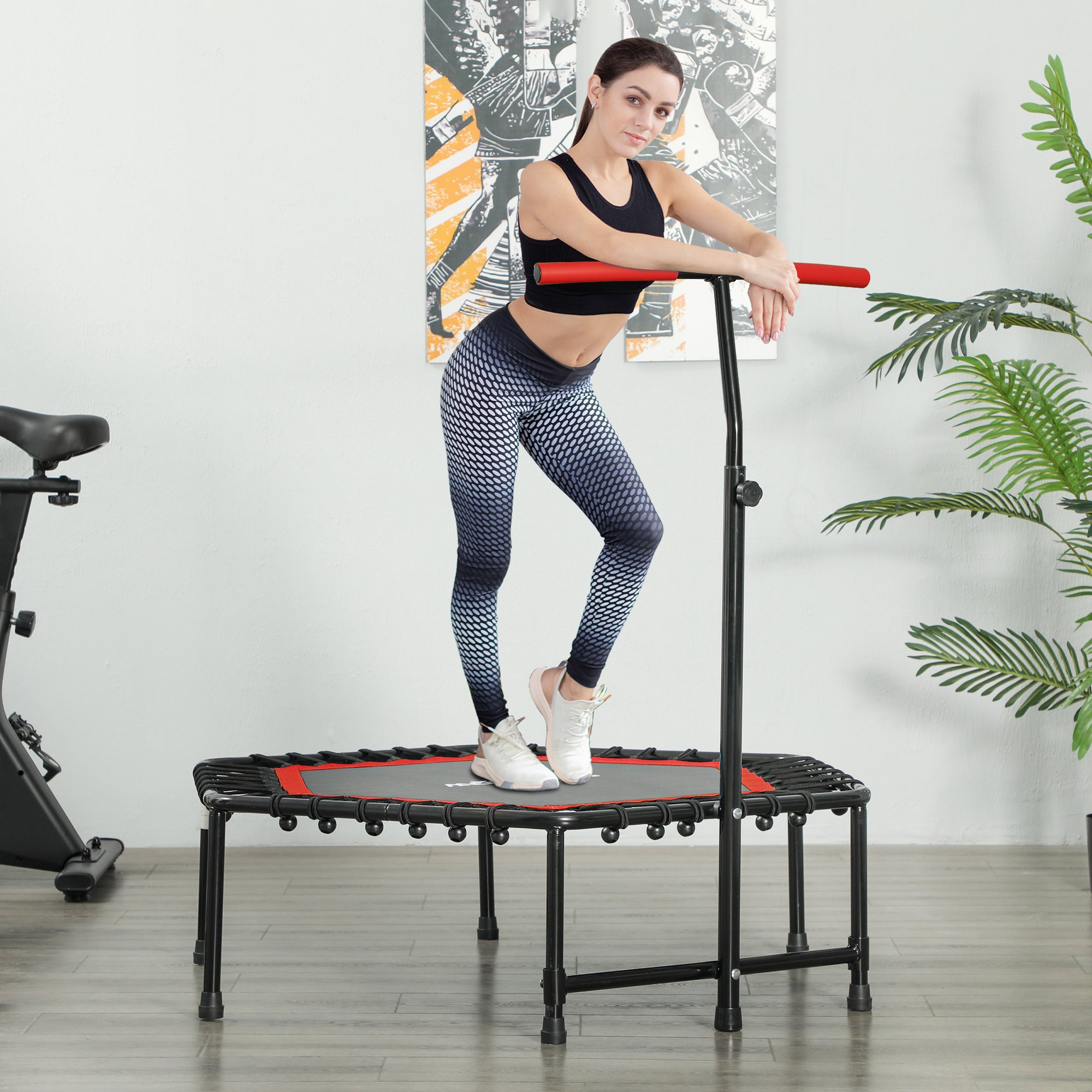 Mujer en ropa deportiva se apoya en trampolín con asa en habitación con pintura y planta.