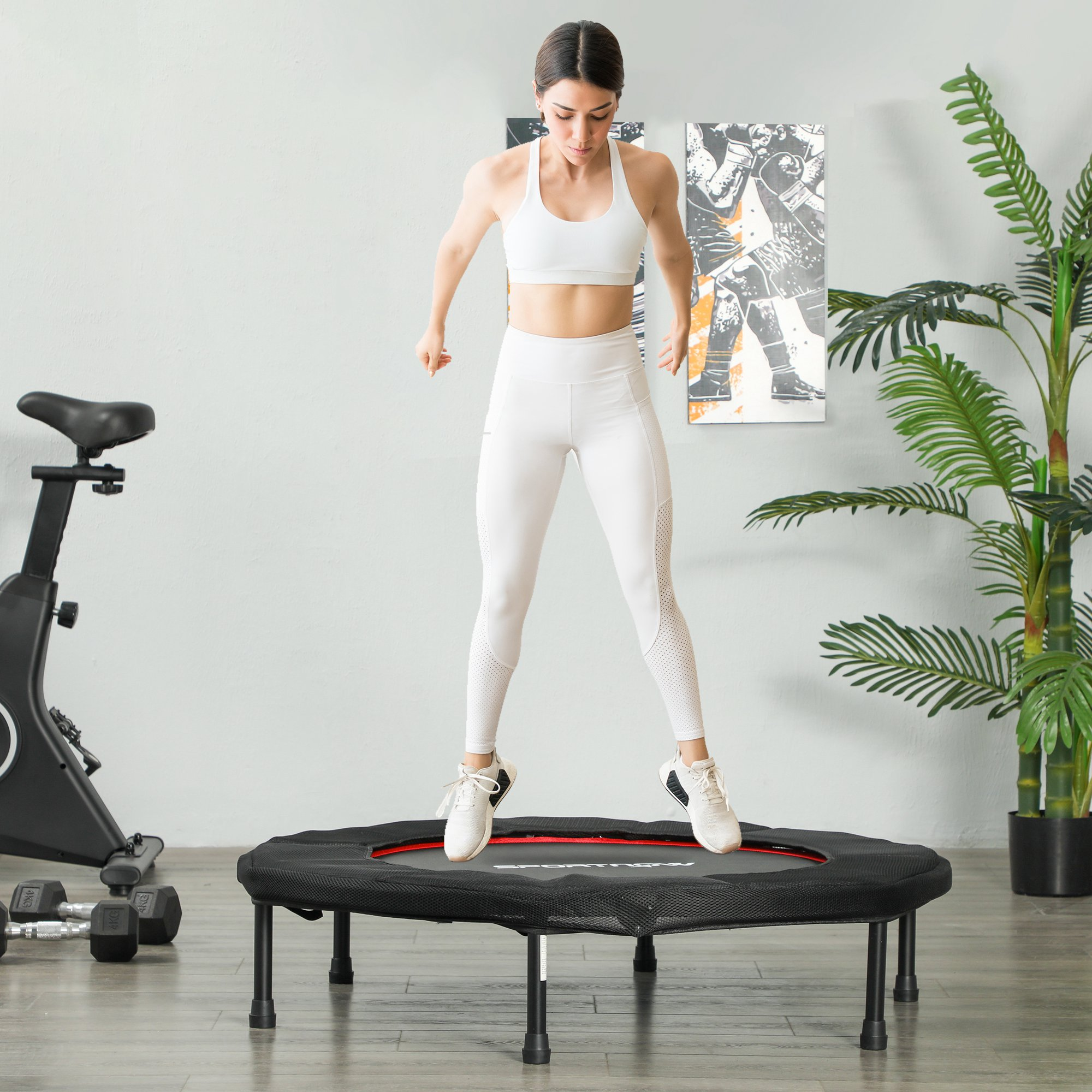 Mujer salta en un trampolín negro. Bicicleta estática y planta visibles en el fondo.