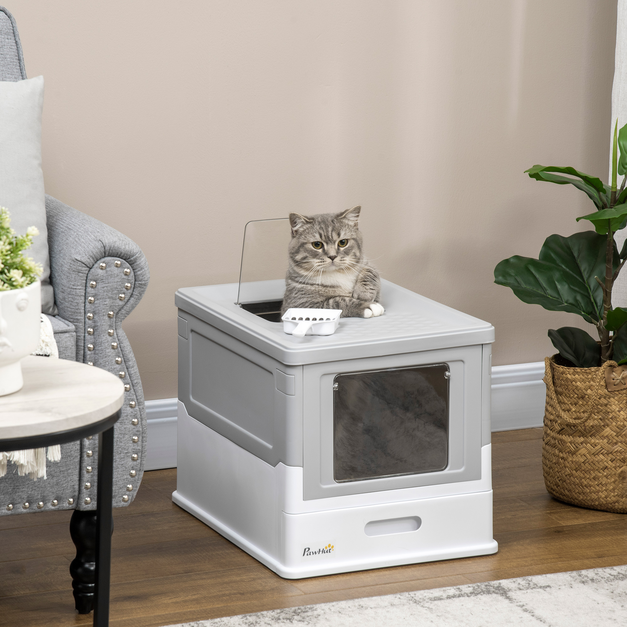 Un gato sentado dentro de una caja de arena gris y blanca en una habitación con mesa y planta.