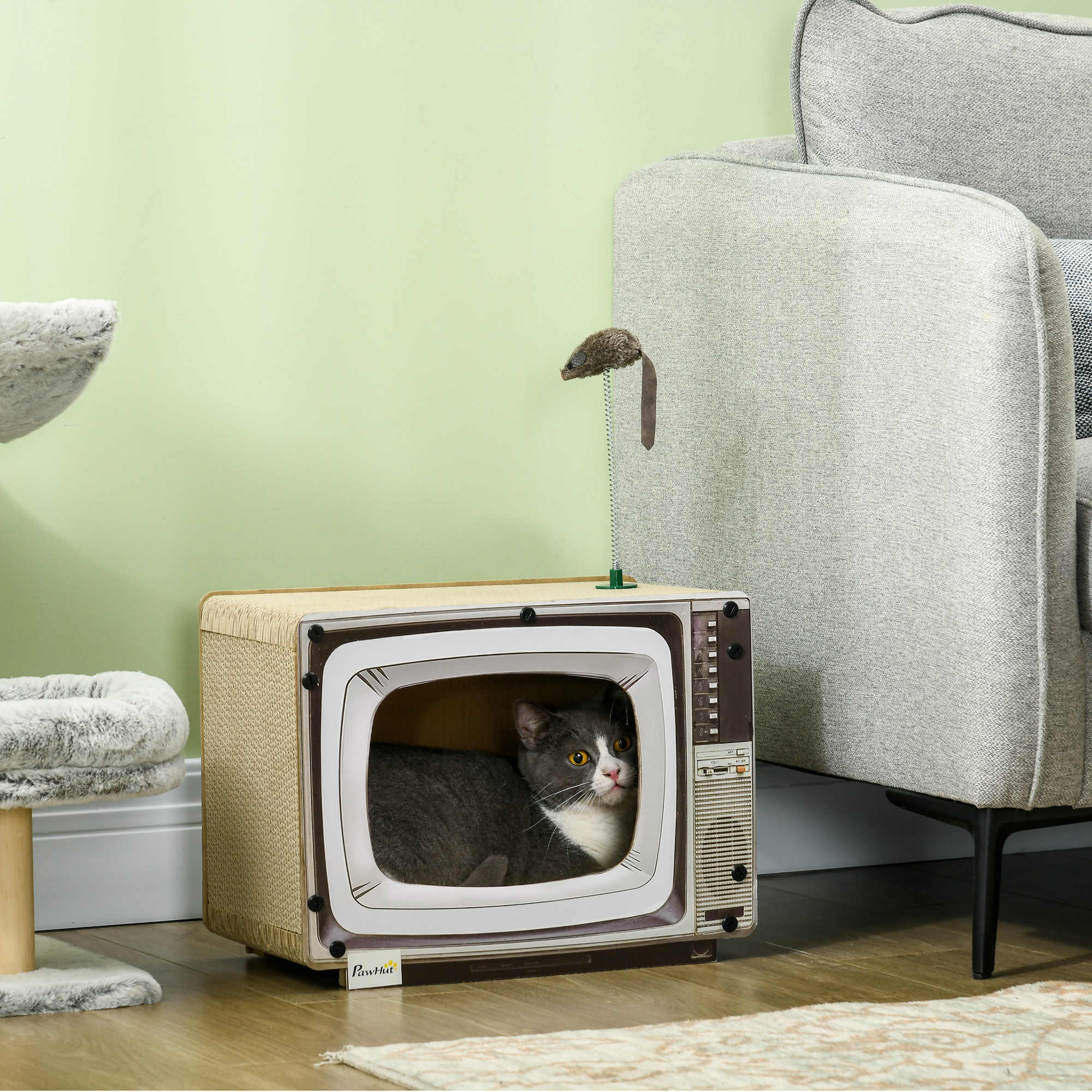 Un gato sentado dentro de una cama para gatos con forma de televisor antiguo con un ratón de juguete encima, en una pared verde.