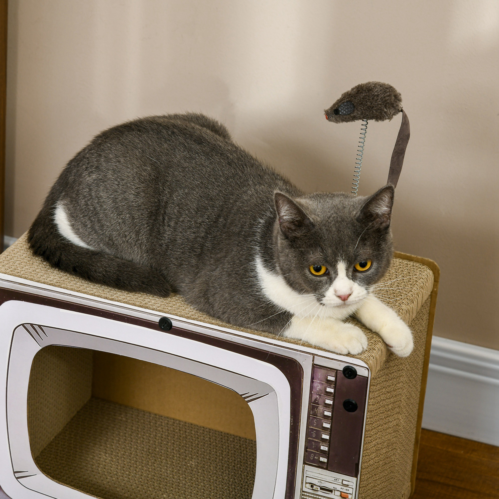 Un gato gris y blanco sentado en una casa para gatos con forma de televisor retro, con un ratón de juguete encima.