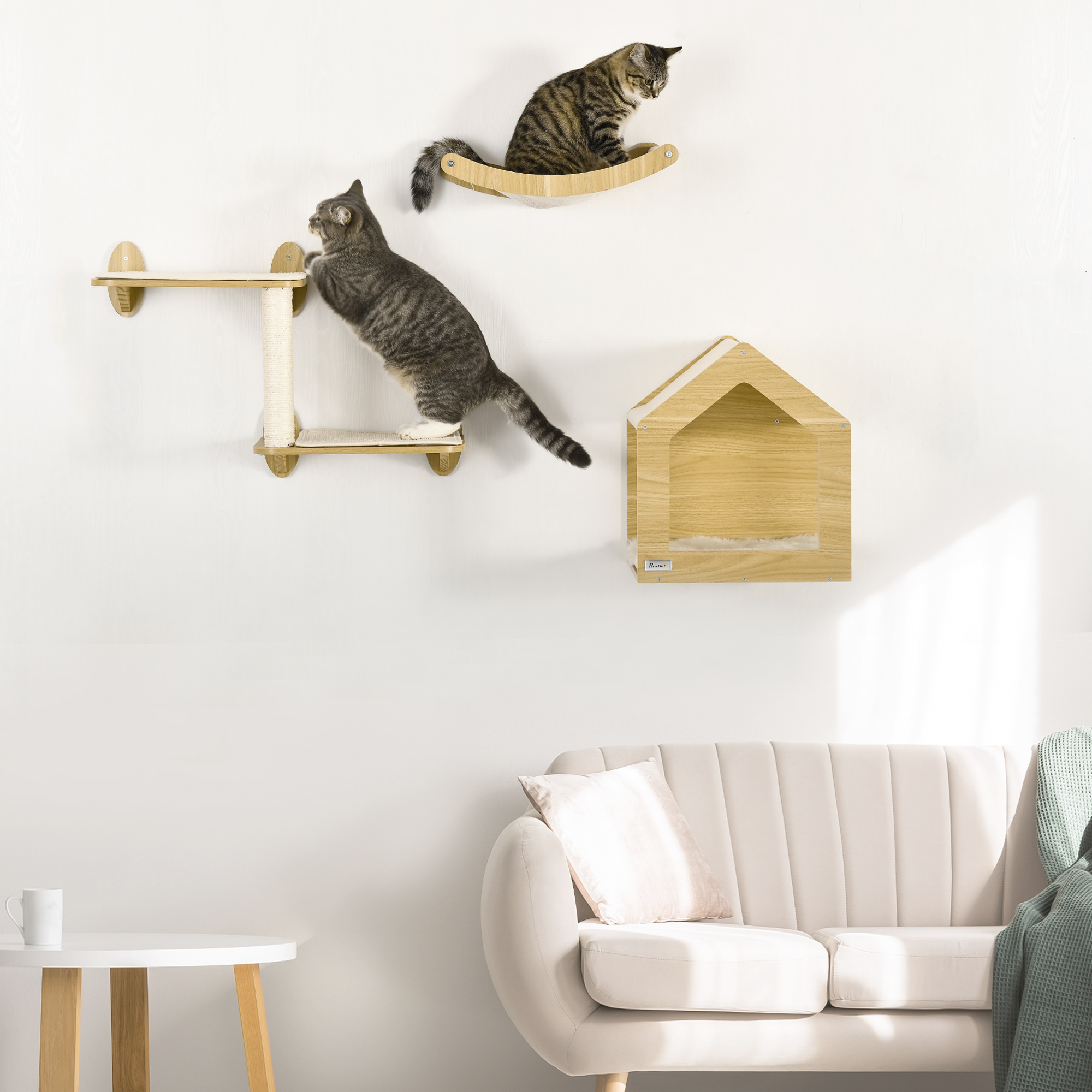 Gatos en árbol para gatos montado en pared. Uno trepa, otro descansa. Sofá beige.