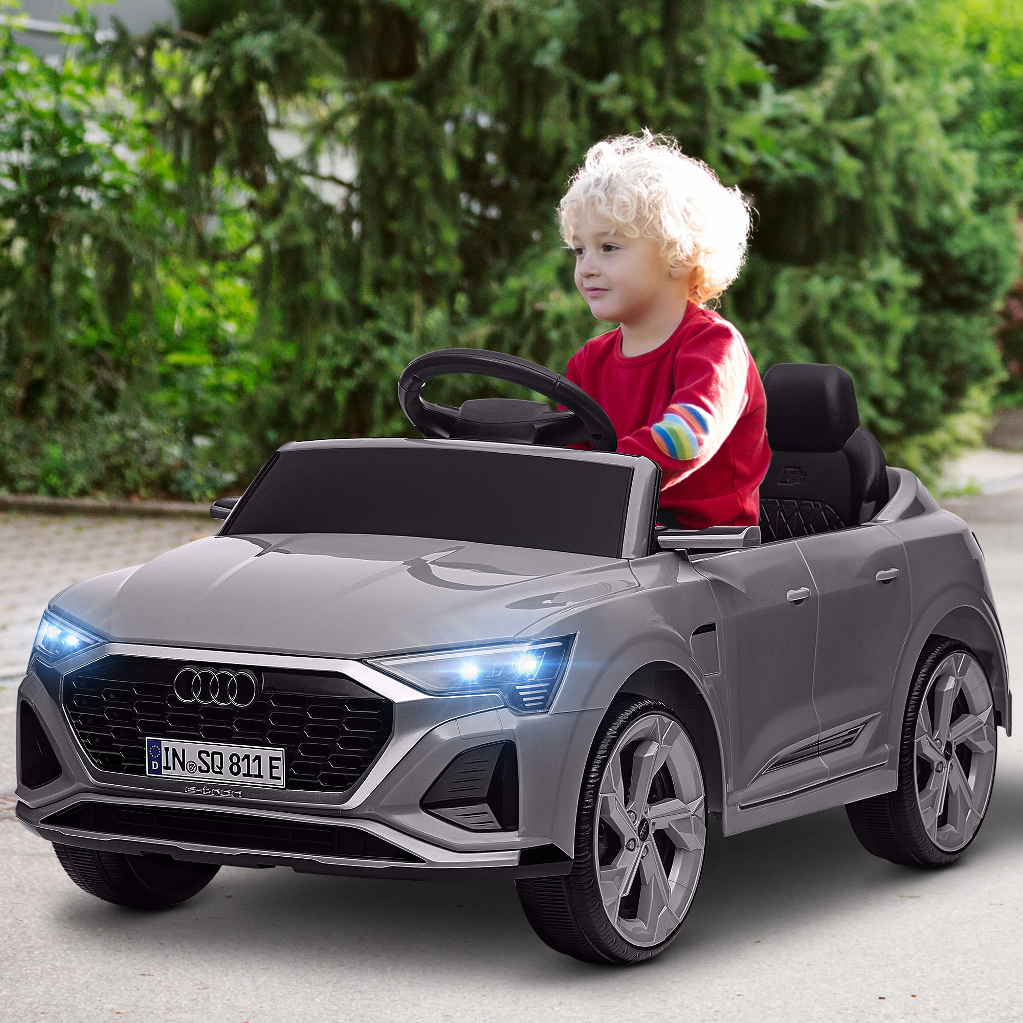 Un niño con camisa roja conduce un coche de juguete Audi Q8 gris en una carretera.