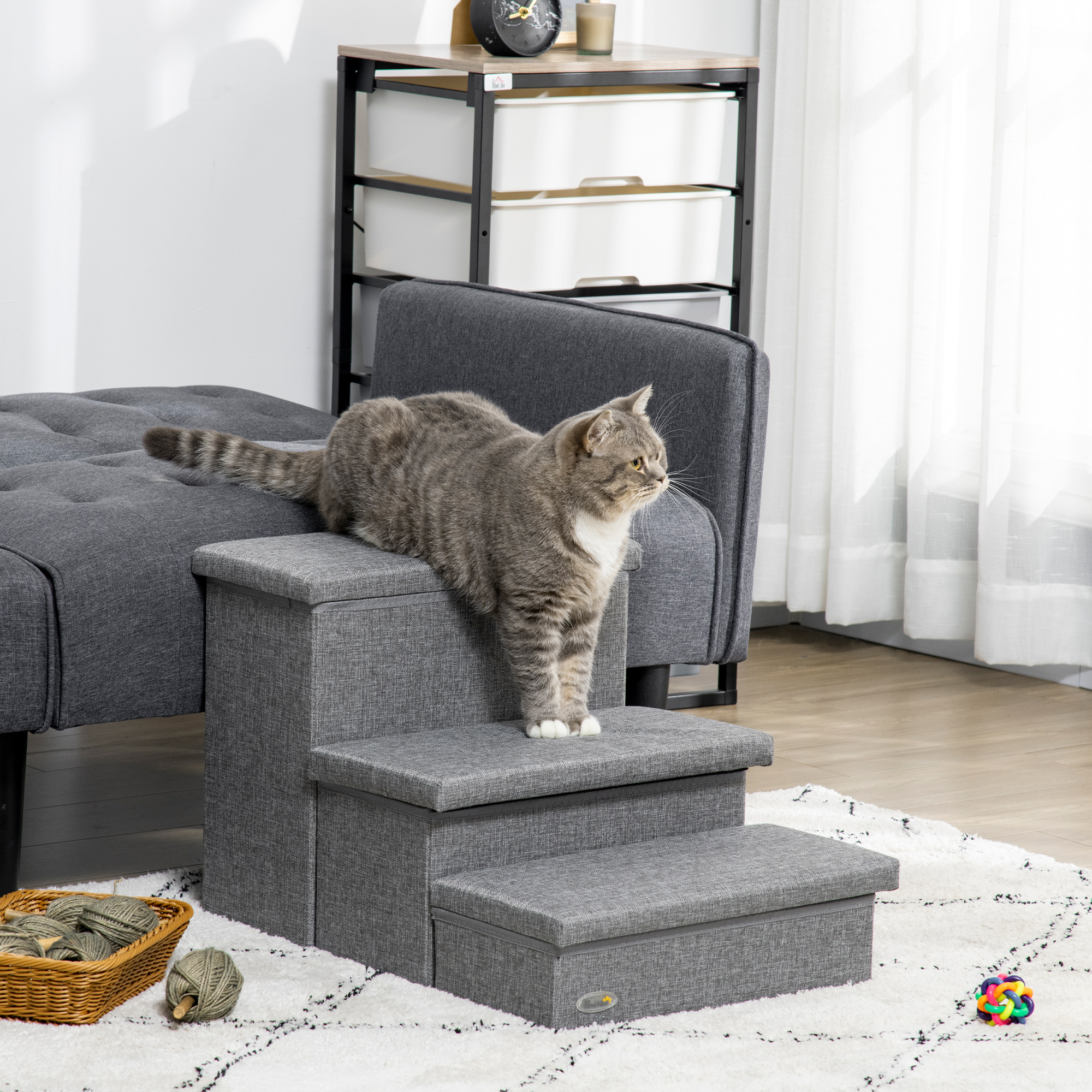 Un gato gris en escalones grises con una cesta de hilo y un sofá detrás.