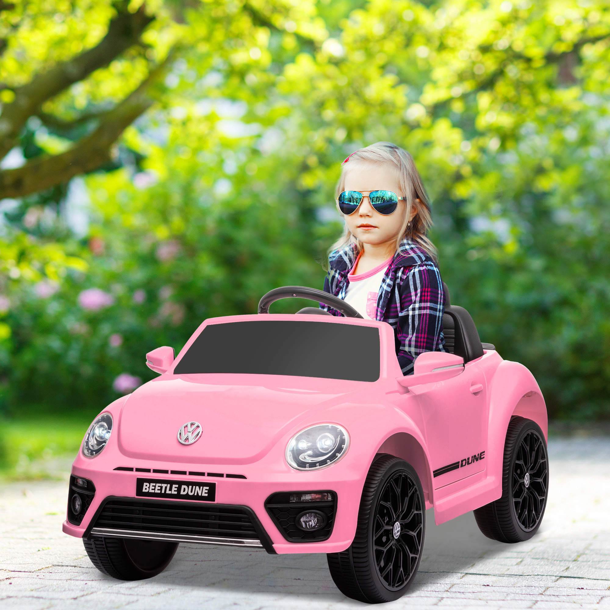 Un niño con gafas de sol conduciendo un coche de juguete rosa en una carretera con un fondo verde y un día soleado.