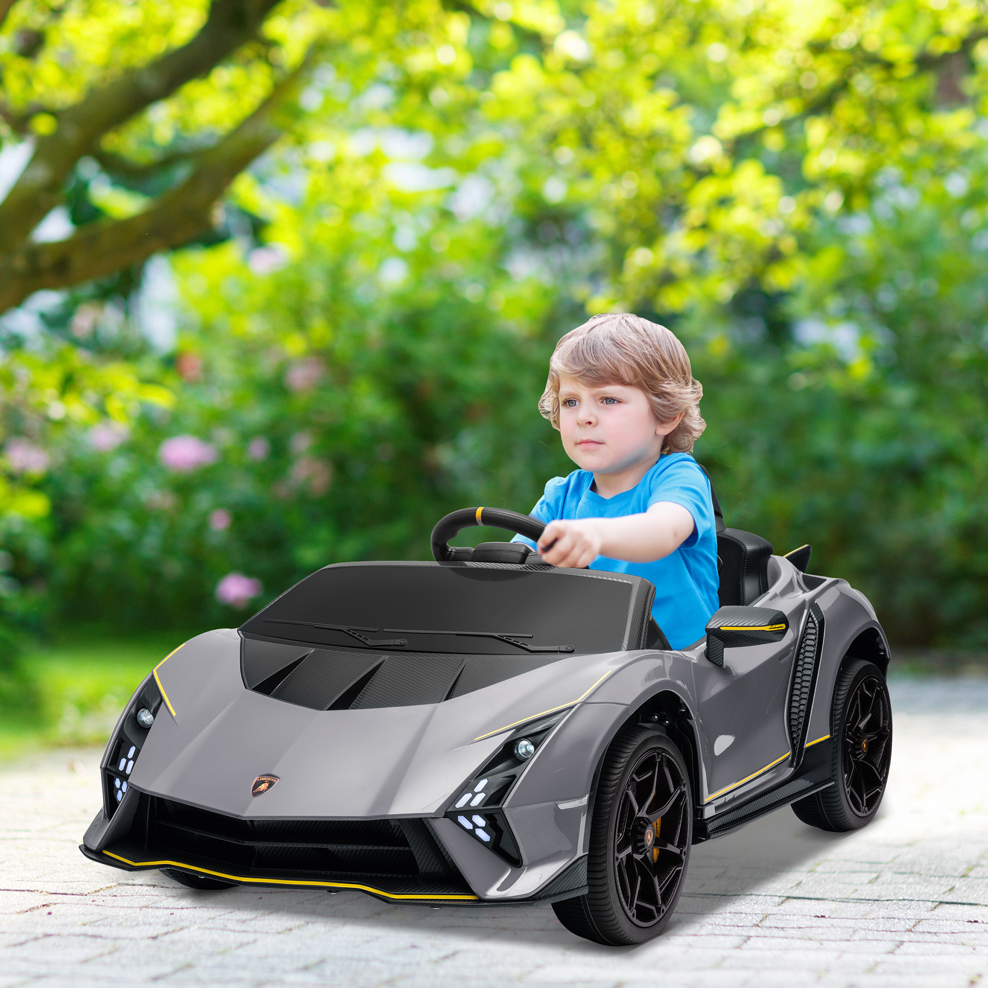 Un niño pequeño conduce un coche de juguete gris en un jardín. El coche es un Lamborghini y el niño lleva una camisa azul.