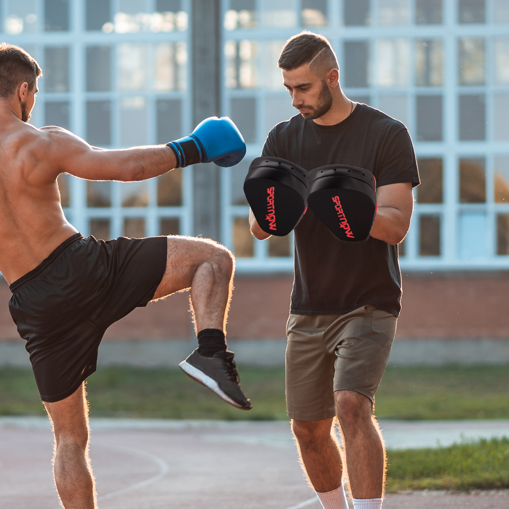 Dos hombres boxeando, uno con guantes azules pateando, uno con almohadillas.