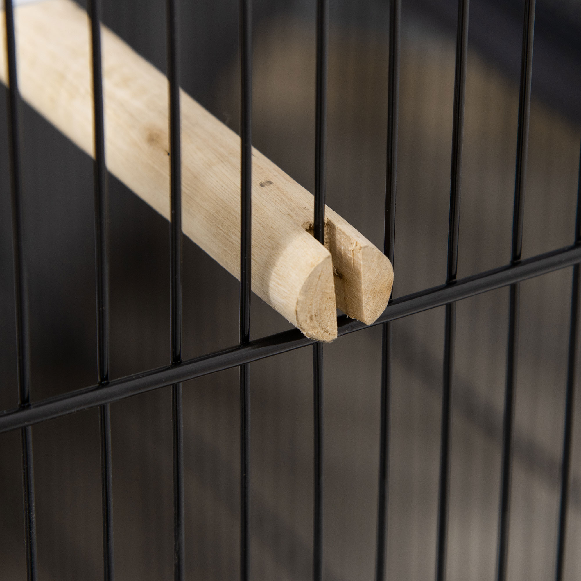 Primer plano de una percha de madera para pájaros en una jaula de alambre negra, mostrando la instalación.