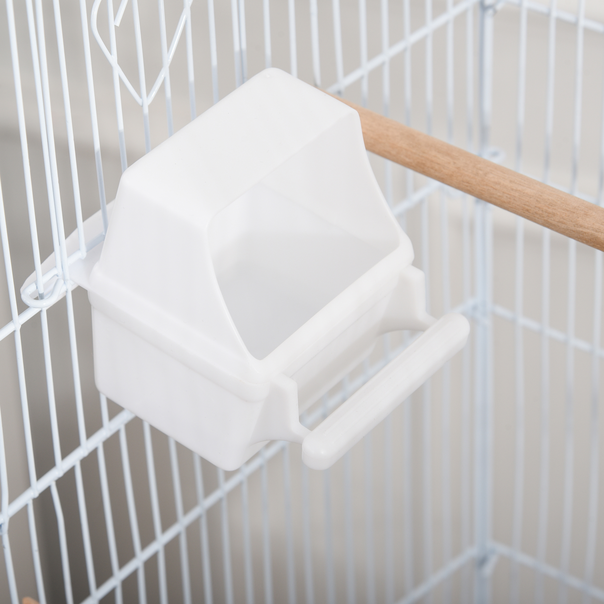 Un comedero blanco para pájaros está adjunto dentro de una jaula. Una percha de madera es visible.