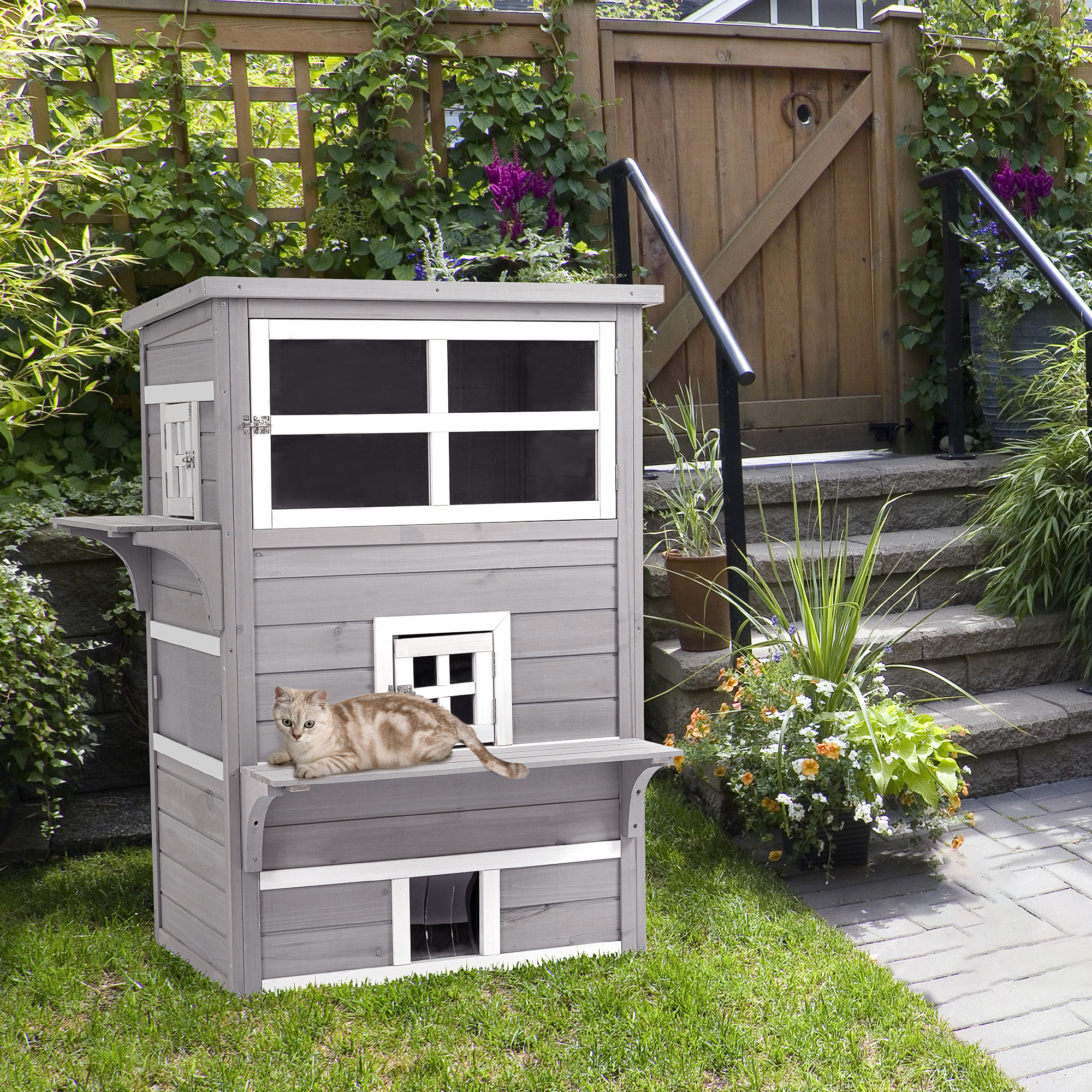 Casa para gatos gris con un gato en la repisa. Hierba verde y valla de madera detrás.
