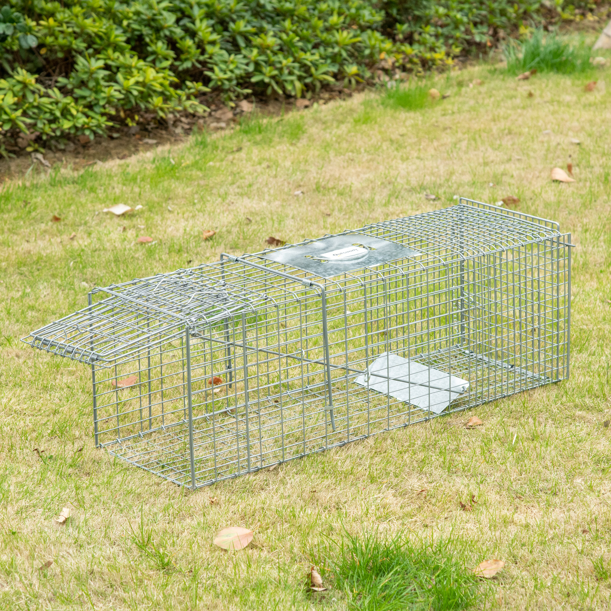 Una trampa metálica para animales vivos está en la hierba. La trampa tiene una puerta abierta.