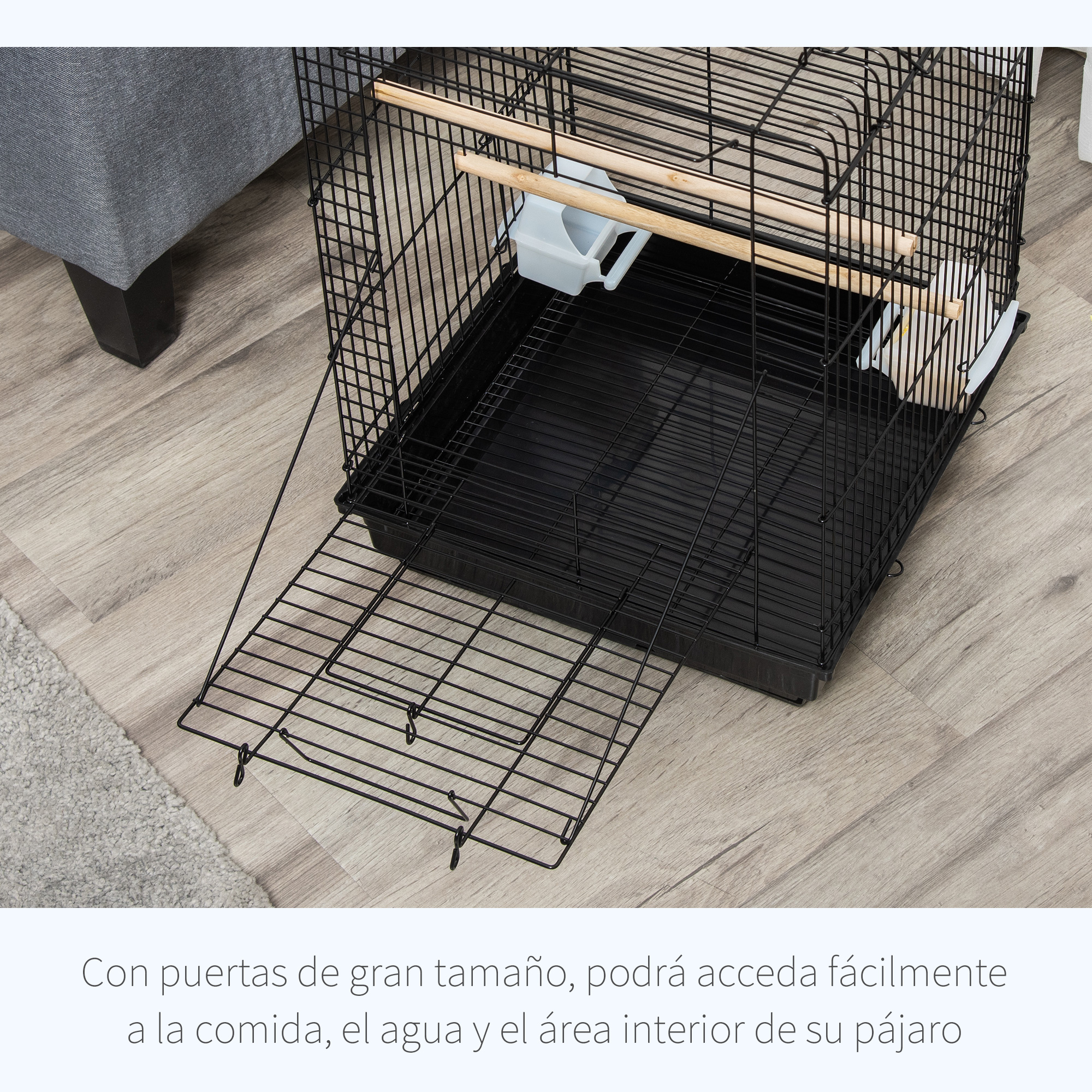 Jaula negra para pájaros en suelo de madera. Puerta entreabierta. Sofá gris visible detrás.