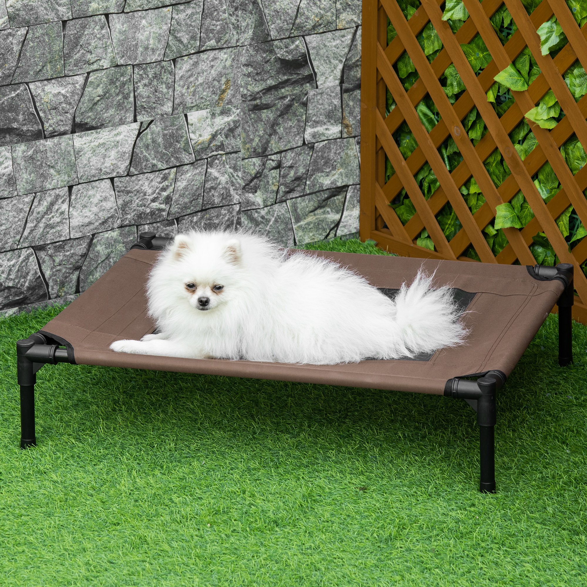 Un perro blanco está acostado en una cama marrón sobre césped verde. Una pared de piedra y una valla de madera al fondo.