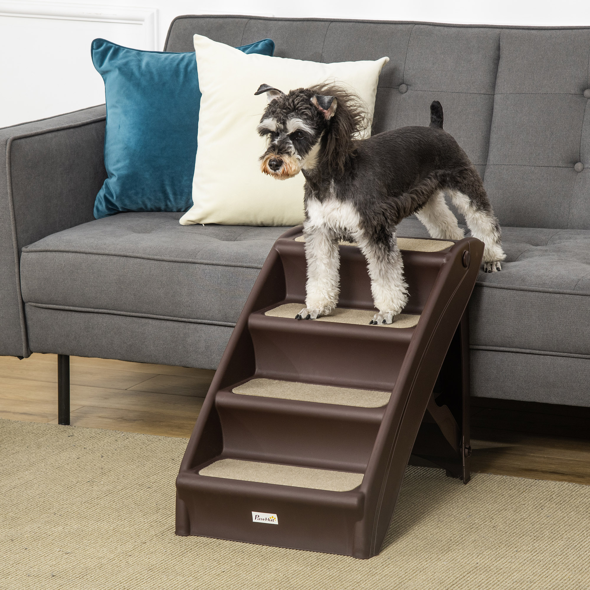 Un perro pequeño en escaleras marrones para mascotas, de pie en el escalón superior, junto a un sofá gris.
