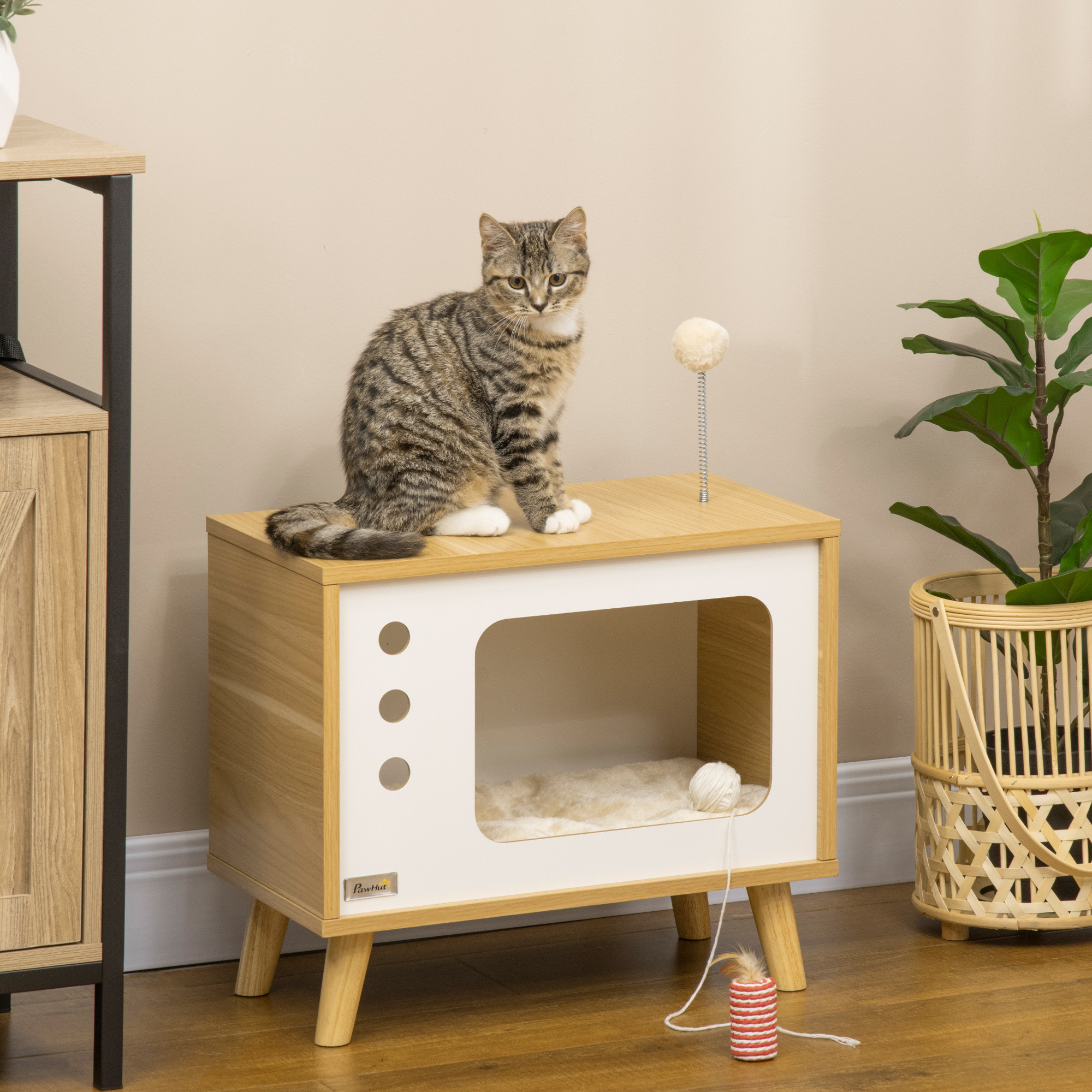 Un gato sentado encima de una casa para gatos que parece un televisor retro. Un juguete está junto a la casa.