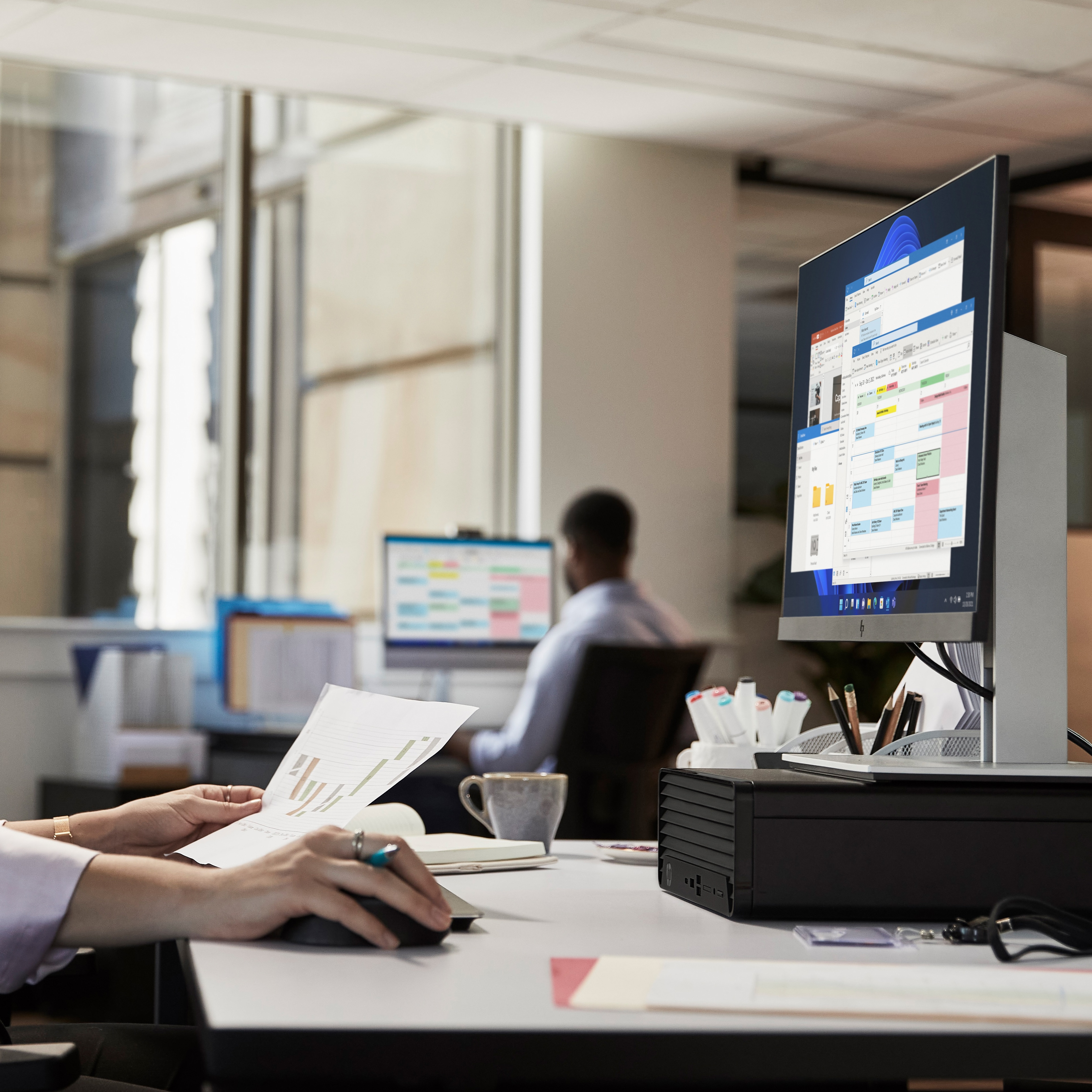 Biurko z komputerem. Osoba trzymająca dokument. Drugi monitor i osoba w tle.