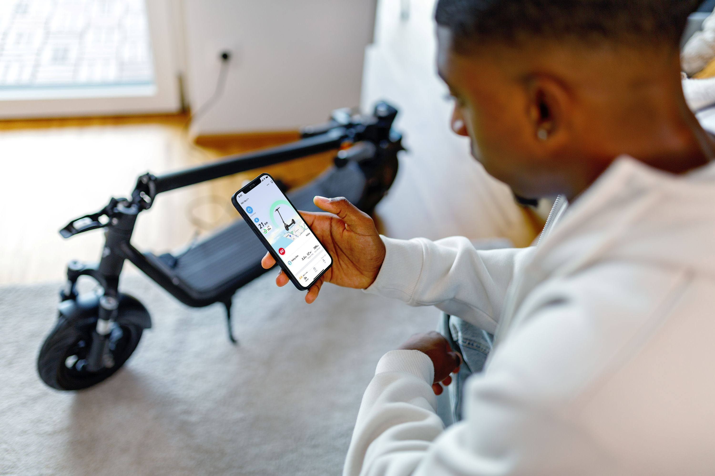 Un hombre sosteniendo un teléfono, mostrando una aplicación sobre un scooter eléctrico. El scooter está en el fondo.