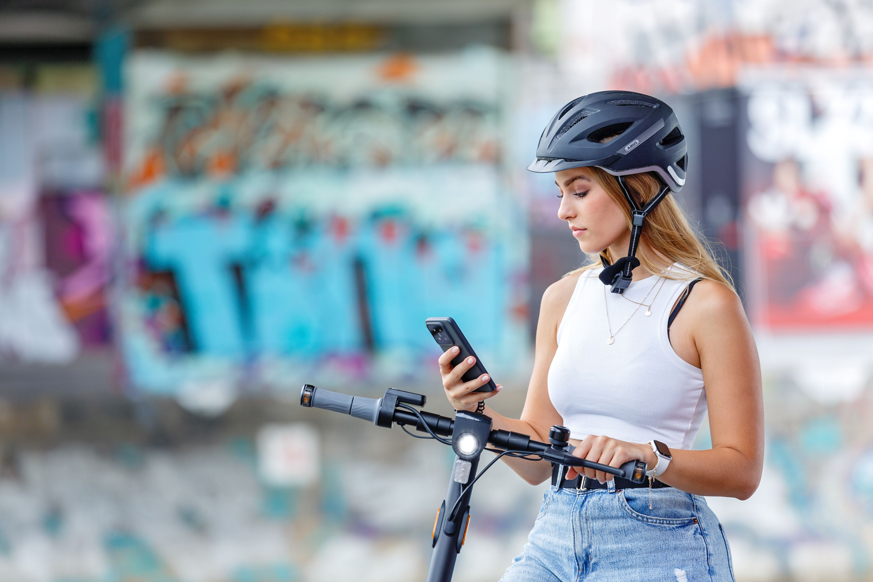 Mujer en patinete mirando el teléfono. Con casco, en un parque, fondo de graffiti.