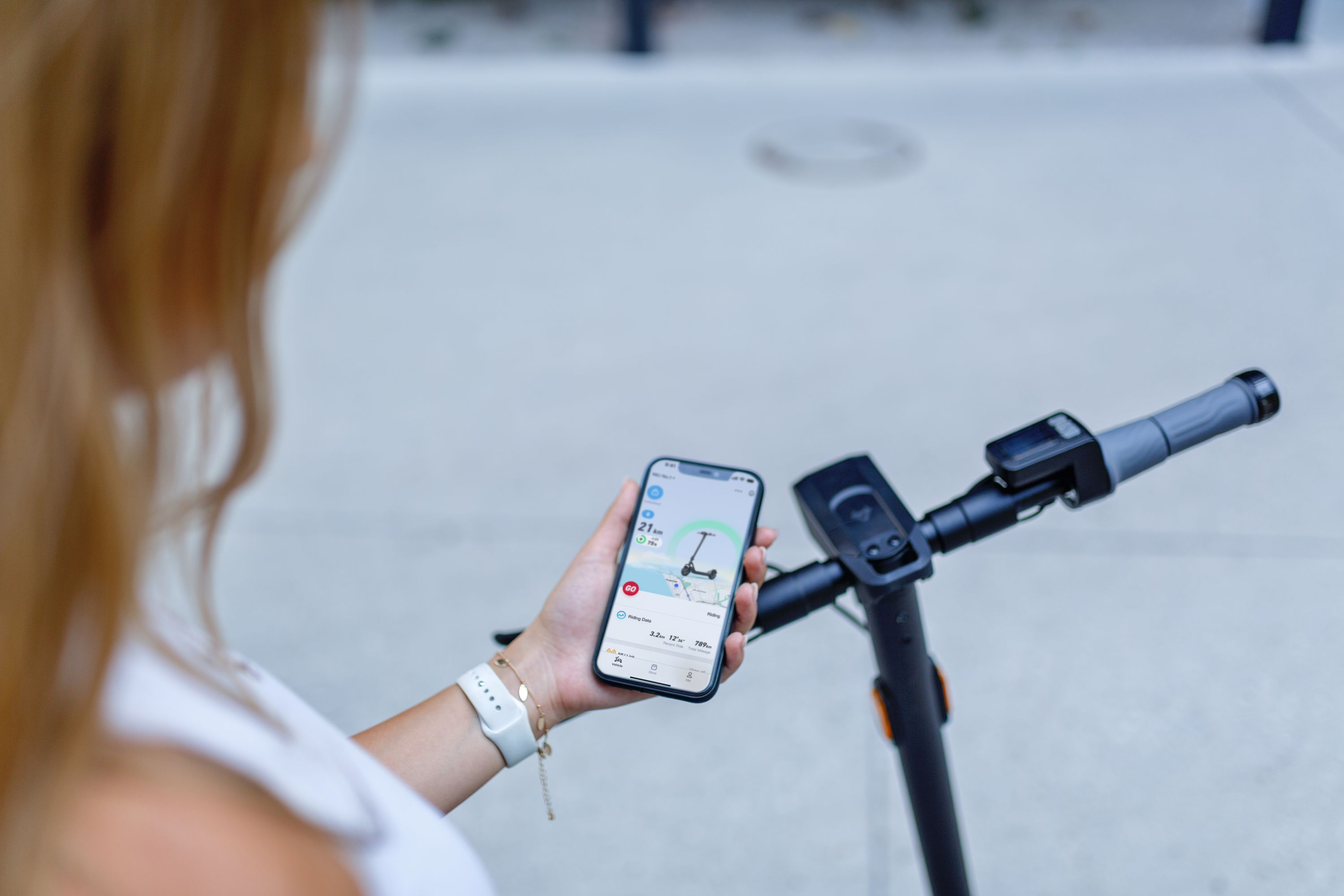 Una mujer sostiene un teléfono que muestra un mapa de patinete eléctrico. El patinete y el manillar son negros.