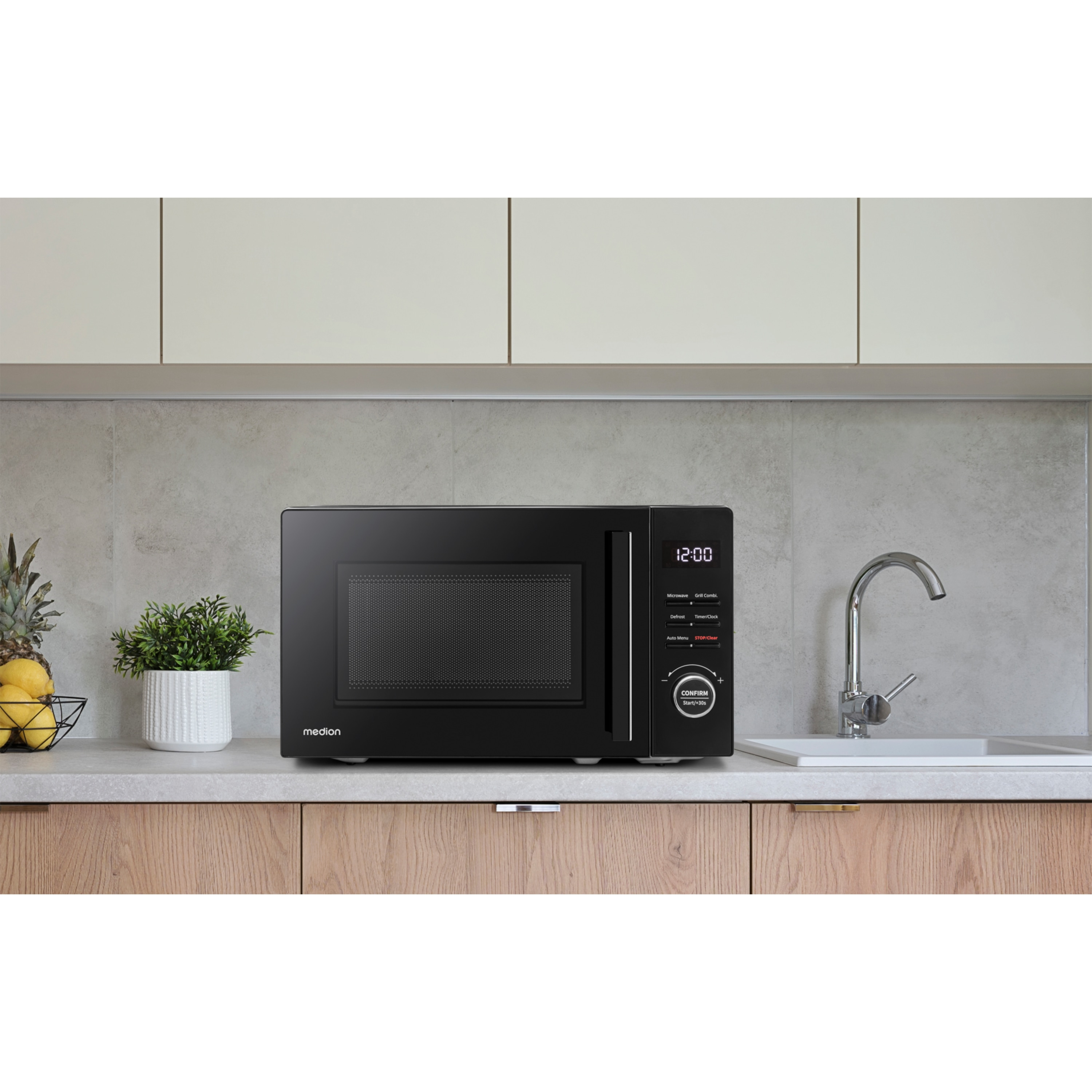 Un horno microondas negro sobre una encimera en una cocina, con una planta, fruta y grifo visibles.