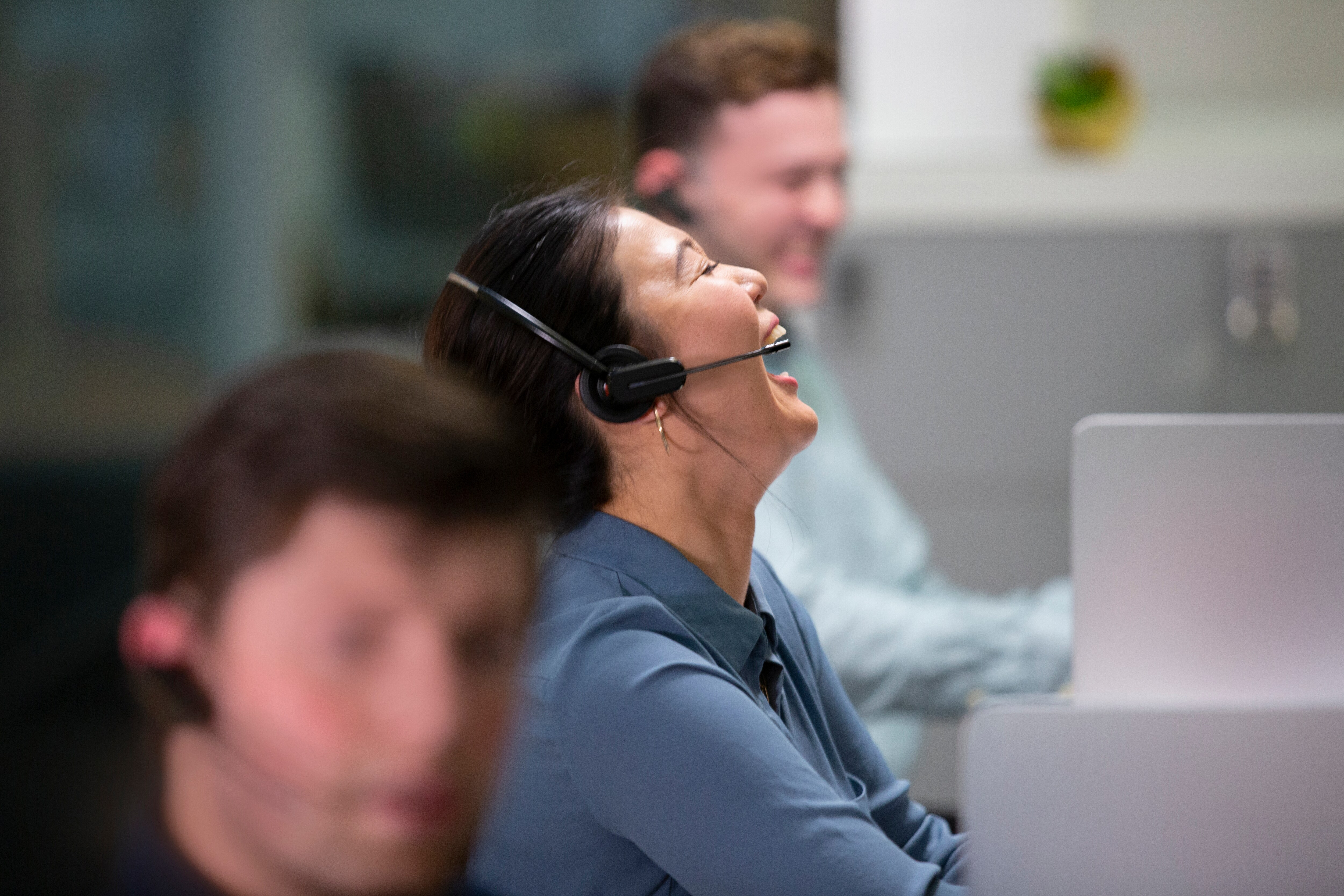 Tre persone in un call center, una donna ride con le cuffie, primo piano sfocato.